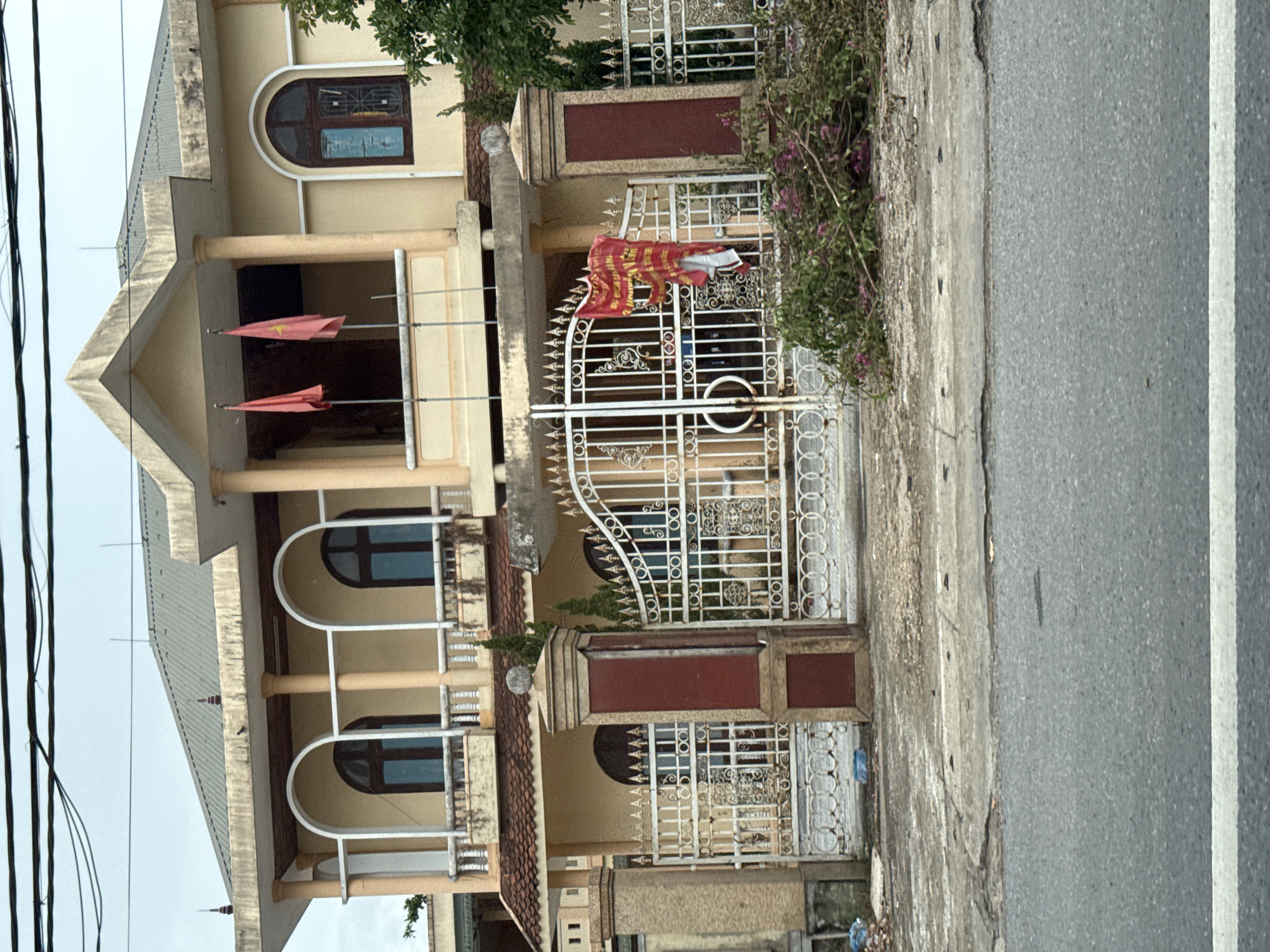 The image shows a large, light yellow, two-story house with a dark gray, corrugated metal roof. The house has a covered balcony on the second floor, supported by beige columns. Two red flags with yellow stars are flying from poles on the balcony. The first floor has an arched entryway with a white metal gate. The gate is decorated with intricate scrollwork and floral motifs. A red banner with yellow writing is hanging from the gate. In front of the gate, there is a pile of green branches with pink flowers. To the left of the gate, there is a beige stone pillar and to the right, a similar pillar with red accents. The house is situated on a street with a gray asphalt road. The sky is overcast and appears to be a light gray. The location is specified as Triệu Phong Commune, Vietnam.