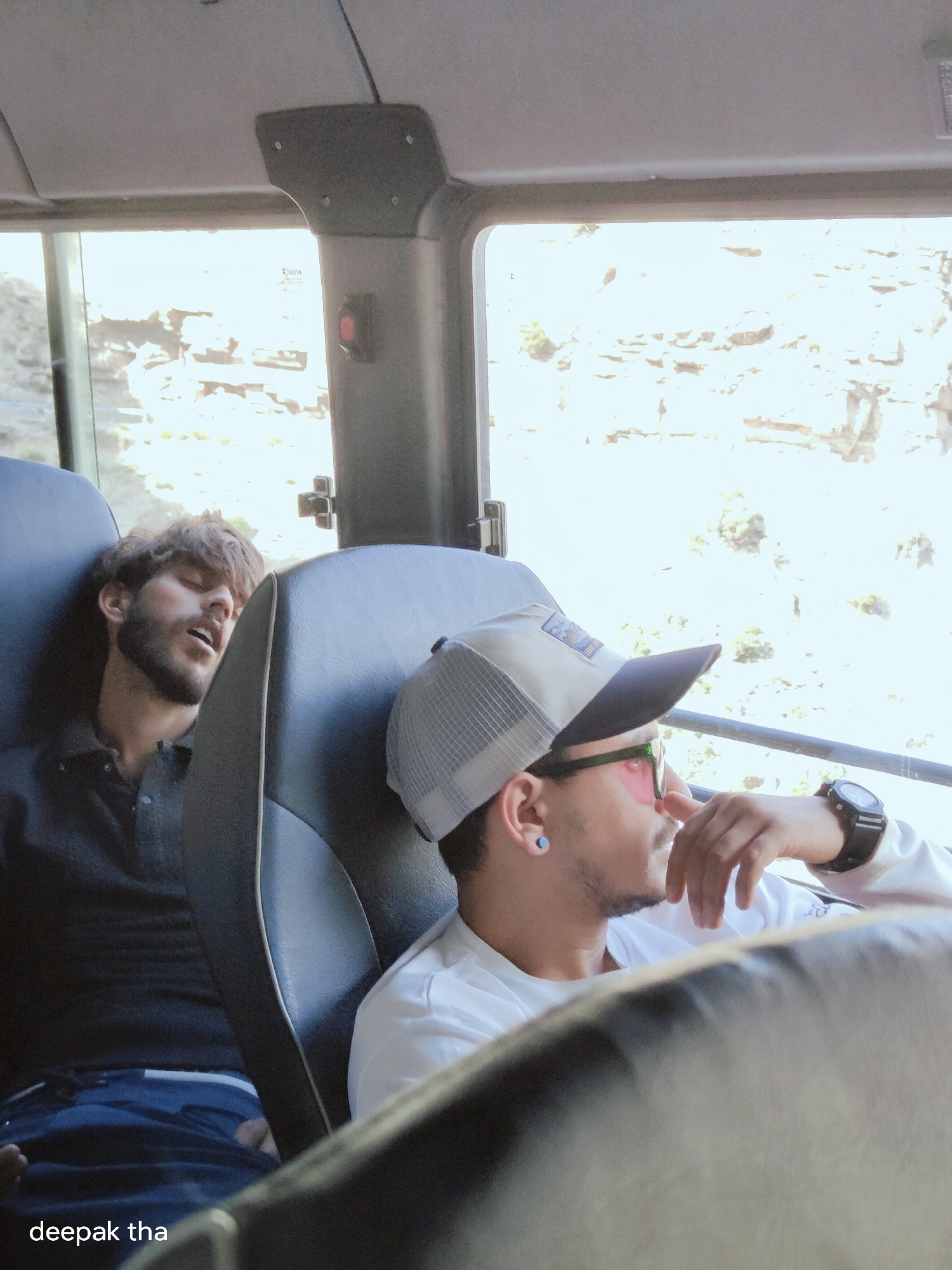 Two men are sitting in a bus. The man on the left is asleep, leaning his head against the seat. He has a beard and is wearing a dark shirt. The man on the right is awake and looking out the window. He is wearing a baseball cap and glasses, and has a watch on his wrist. The bus appears to be traveling through a scenic, mountainous area. There is text at the bottom of the image that reads "deepak tha". It is not possible to determine the exact location within Stingri, India, based on the provided image.