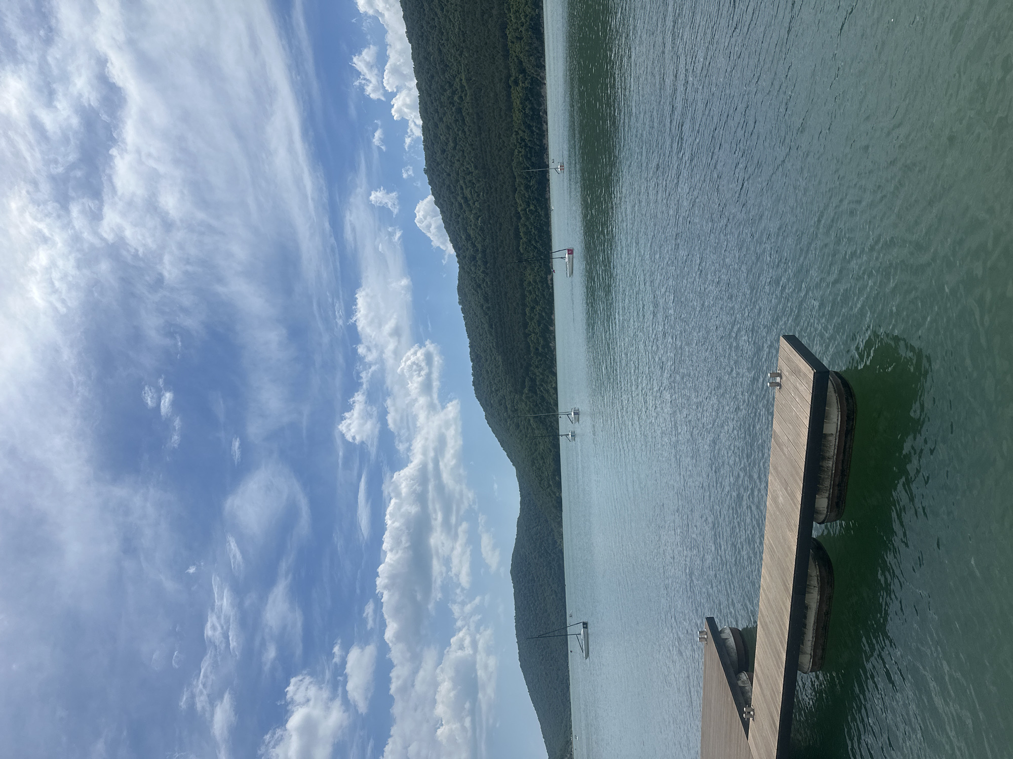 The image depicts a scenic view of a body of water, likely Abrau Lake, in Abrau-Dyurso, Russia. In the foreground, a wooden pier extends into the tranquil blue water. The pier is constructed with wooden planks and supported by buoys. Several sailboats are visible on the lake, with their masts piercing the sky. In the background, lush green hills covered in trees rise majestically, forming a natural backdrop to the serene landscape. The sky above is a brilliant blue, adorned with fluffy white clouds, suggesting a clear and pleasant day.