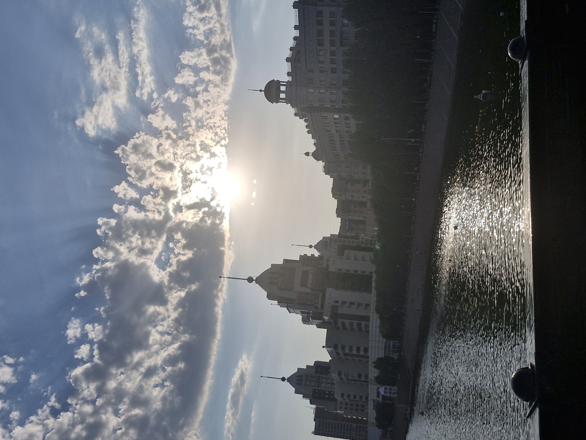 The image captures a view of the Ishim River in Astana, Kazakhstan, with a distinctive building on the right and a series of layered architectural structures on the left, characterized by stepped facades and spire-like elements. The sun is setting, casting a bright glare that filters through scattered clouds, creating rays of light. The river's surface reflects the sunlight, appearing textured and shimmering. Along the riverbank, a line of trees separates the water from the buildings. In the foreground, a dark silhouette of a railing with decorative spherical finials is visible. The overall scene depicts an urban landscape during the golden hour.
