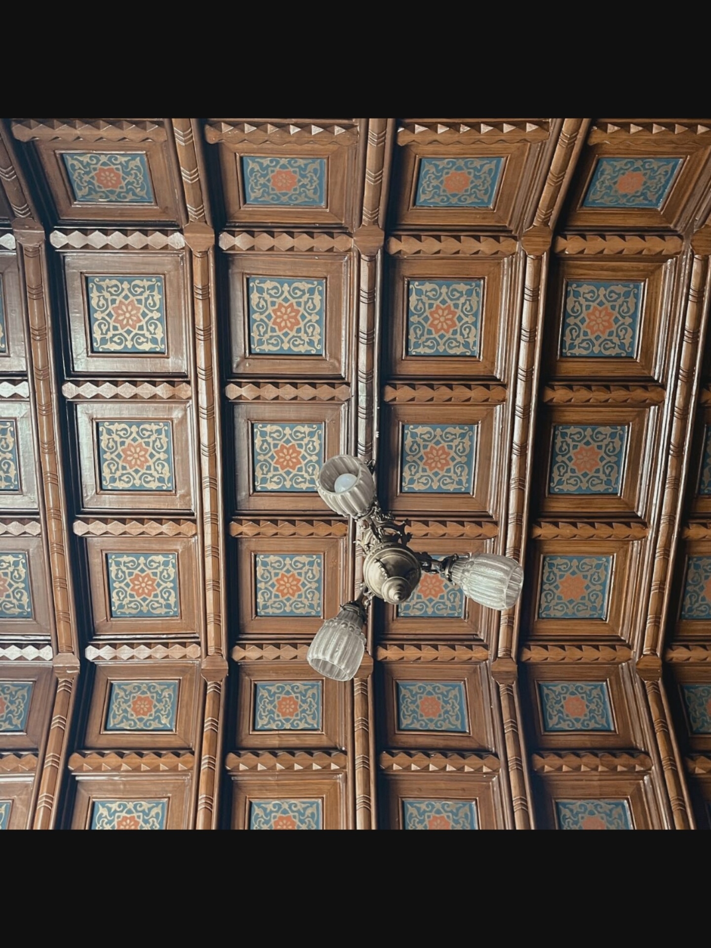This is a low-angle, full shot of an ornate wooden coffered ceiling in Saint-Août, France. The ceiling is composed of square panels with intricate geometric patterns and floral motifs in shades of orange, blue, and gold. A vintage chandelier with three glass shades hangs from the center of the ceiling. The chandelier is made of brass and has a detailed, decorative arm. The lighting in the scene is dim, suggesting an indoor environment. The image focuses on the architectural details of the ceiling and the decorative light fixture.
