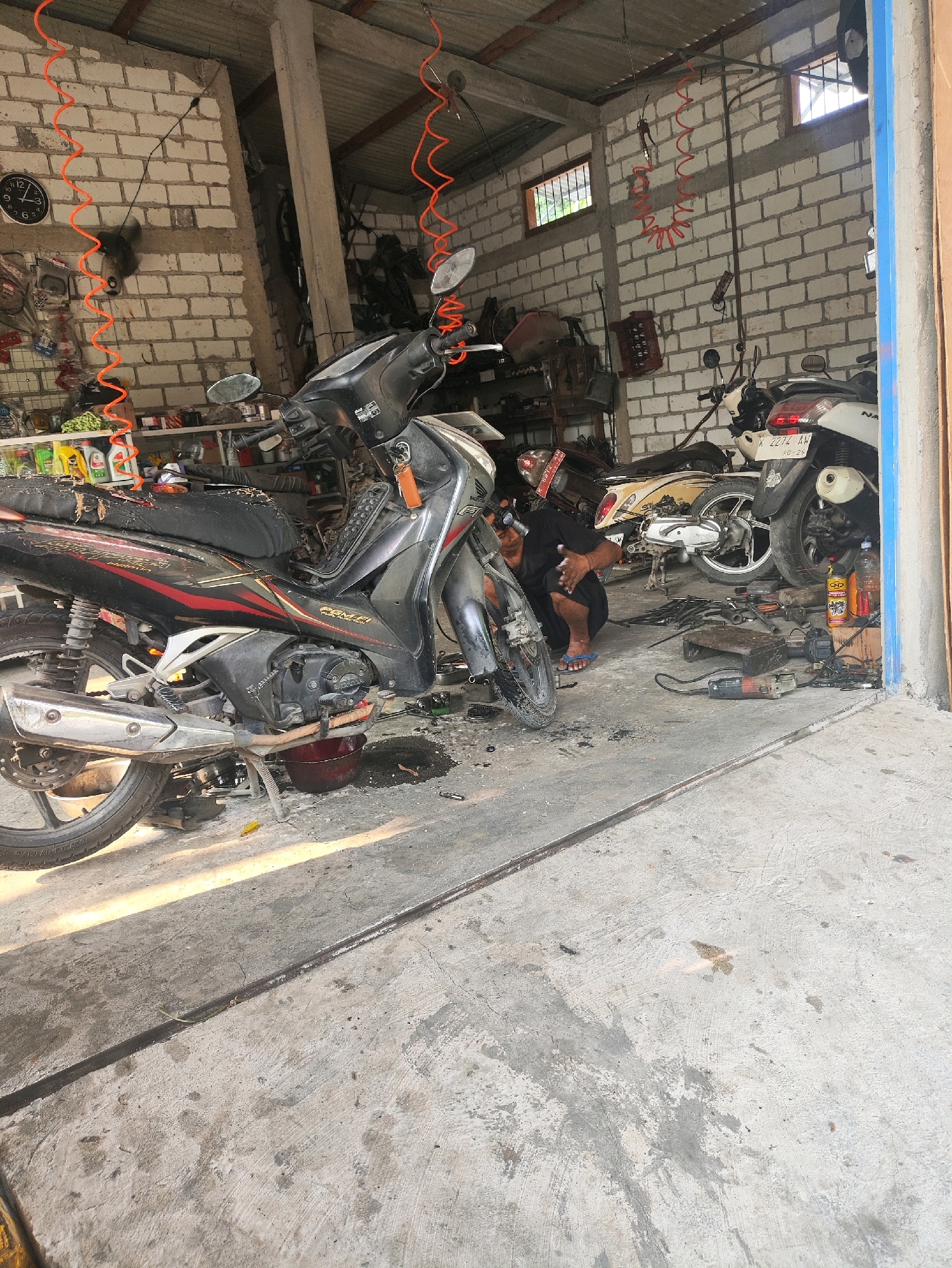 The image depicts the interior of a motorcycle repair shop located in Ngadem, Indonesia. The scene is cluttered with various motorcycles, tools, and repair equipment. In the foreground, a black and red motorcycle is parked on a concrete floor, with an oil pan placed beneath it, suggesting it is undergoing maintenance. A person wearing a black shirt and dark pants is kneeling near the motorcycle, appearing to be working on it. To the right, another motorcycle, a white scooter, is also visible. The walls of the shop are constructed from white cinder blocks. Orange coiled hoses hang from the ceiling, likely air hoses for tools. Shelves with various items, including bottles of oil and cleaning supplies, are present in the background. The overall impression is of a busy, working repair shop.