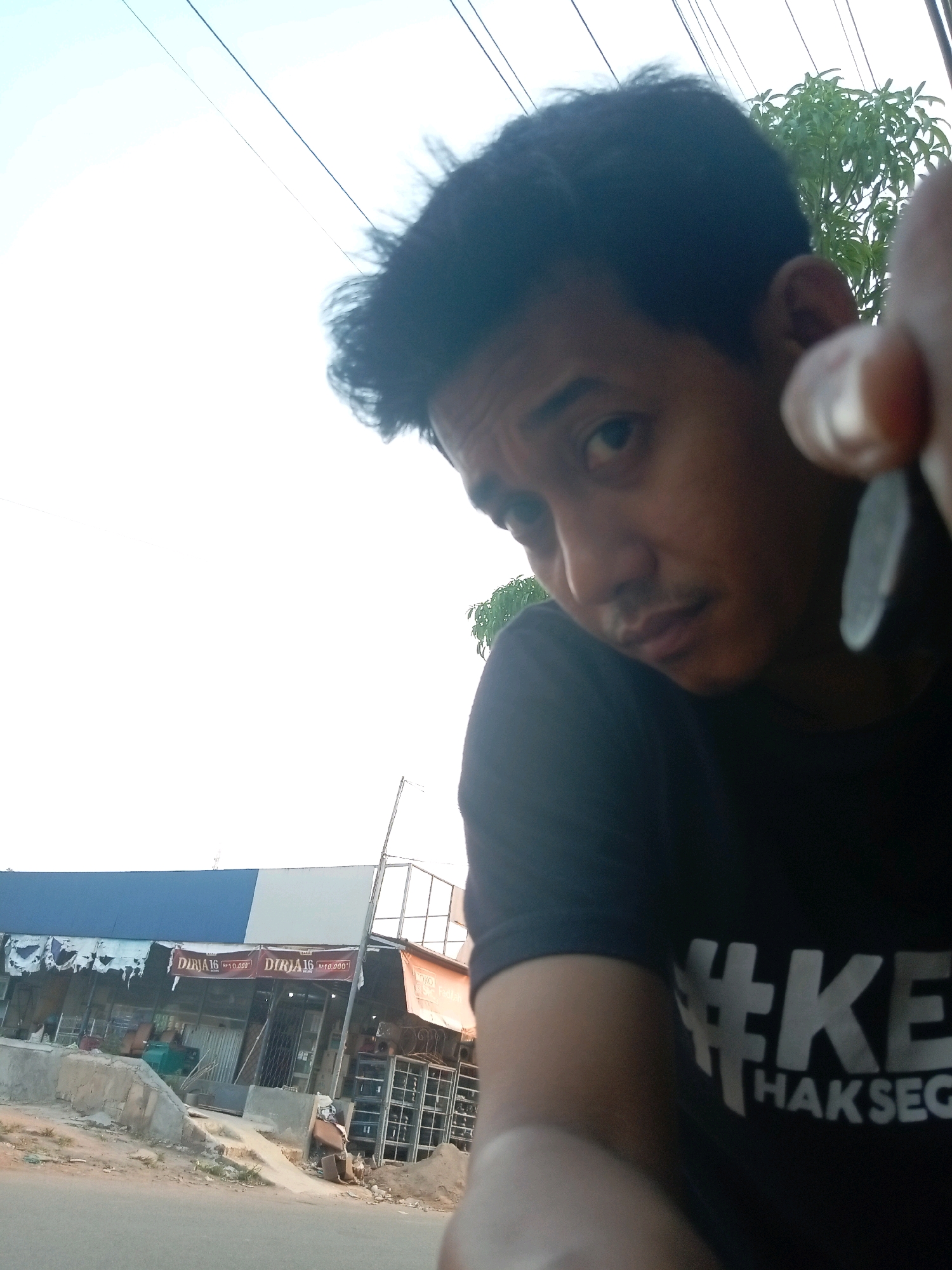 A man, his face partially obscured by his hand holding a coin, is taking a selfie outdoors. He is wearing a black t-shirt with a white hashtag and text that appears to say "#KE HAKSEG". In the background, there is a row of storefronts with blue and white signage. One sign visible reads "DIRJA16 RP10.000". The area appears to be an urban setting with a paved road in the foreground and some vegetation and power lines visible. The image has a slightly tilted perspective.