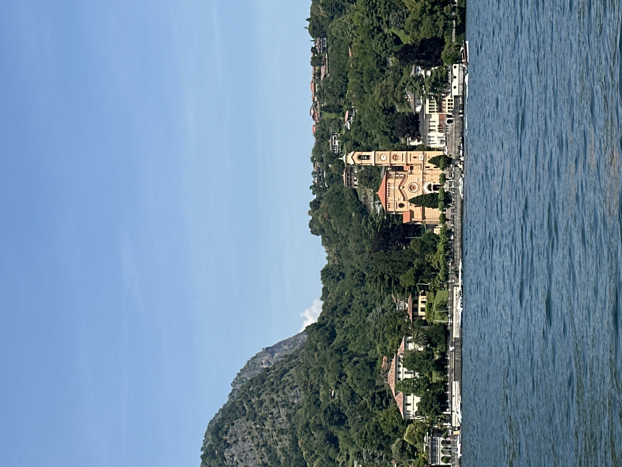 This is a full-frame, eye-level, outdoor, horizontal photograph taken from a body of water, likely Lake Como in Tremezzina, Italy. The scene depicts a shoreline dotted with buildings and lush green trees. In the center-left of the frame, a prominent bell tower and a Romanesque-style church with a red tiled roof stand out. The church is painted a light orange-yellow and has circular windows. To the left of the church, a large white villa with arched windows and a terracotta roof is visible, partially obscured by trees. Further left, other residential buildings with terracotta roofs are nestled along the shore. A steep, rocky mountain covered in dense green trees dominates the background on the left side of the image, with a lighter rocky face visible near its peak. The right side of the background shows a hillside covered with more trees and scattered houses with red roofs. The foreground is filled with deep blue, choppy water, suggesting a breezy day. A white motorboat with two visible occupants is cutting through the water on the right side, creating a wake. Several other smaller boats are docked or moving slowly along the shoreline. The sky is a clear, light blue with a few wispy white clouds on the horizon.