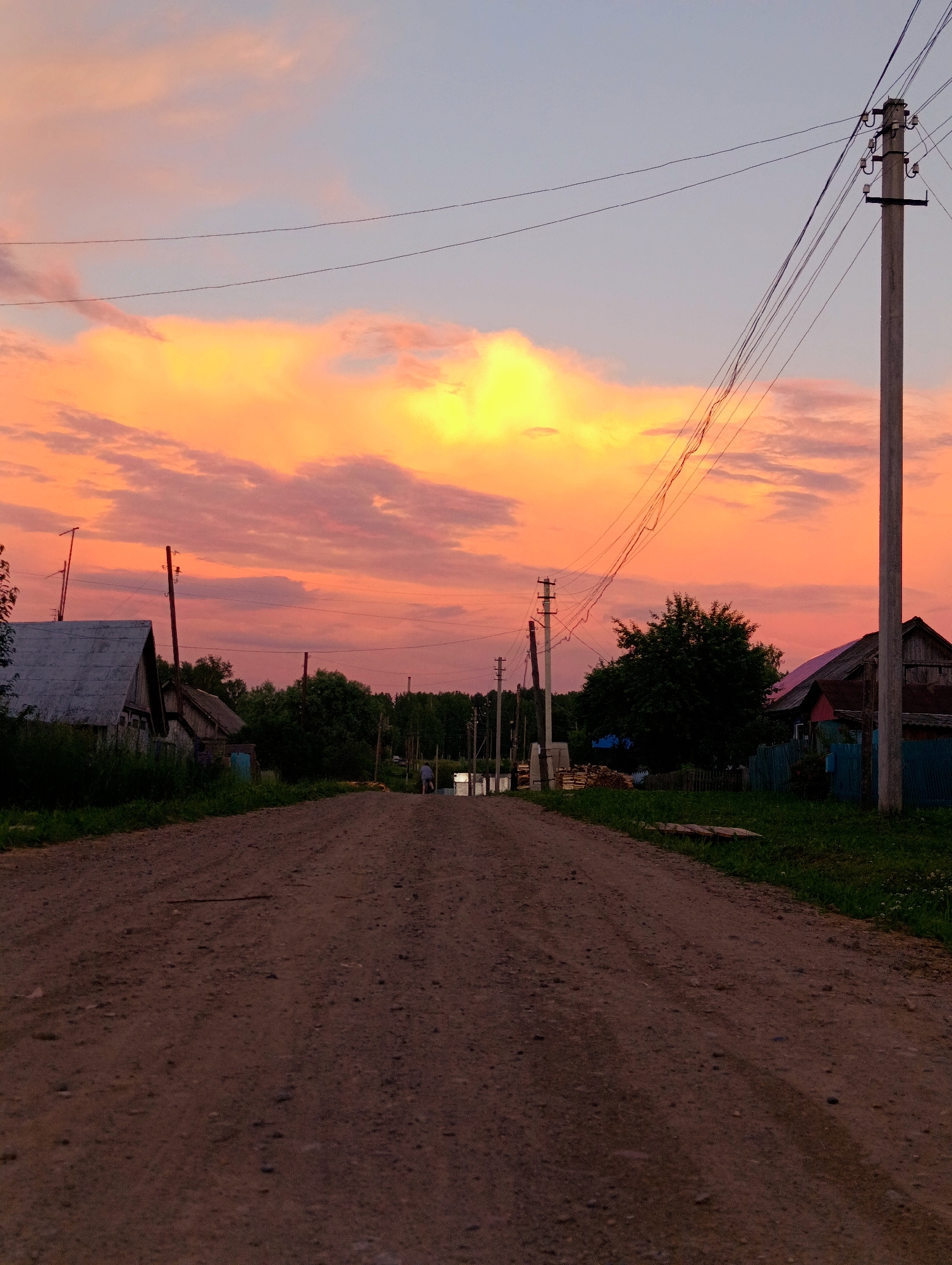 The image captures a rural landscape in the village of Zipunovo, Russia, during a vibrant sunset. A dirt road stretches into the distance, flanked by sparse vegetation and several rustic wooden houses. Power lines traverse the scene, supported by utility poles, with one prominent pole in the foreground on the right. The sky is a dramatic spectacle of orange, pink, and yellow clouds, illuminated by the setting sun, against a soft blue backdrop. A lone figure can be seen in the distance, walking along the road. The overall impression is one of quiet rural life against a breathtaking natural backdrop.