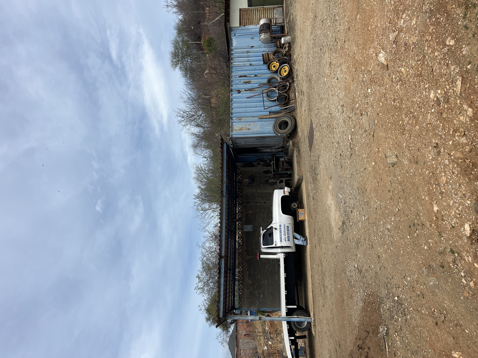 In the city of Juan Griego, Venezuela, a tow truck is parked under a blue metal awning in an outdoor workshop. The truck is white and has a flatbed. A person wearing blue jeans and a dark shirt stands next to the driver's side door of the tow truck, appearing to be working on the vehicle. The workshop also features a large blue shipping container, various tires, and metal rims stacked in front of it. The ground is covered in dirt and gravel. In the background, trees and vegetation are visible on a hillside, with a cloudy sky overhead. The scene depicts an automotive repair or service area in a semi-arid environment.
