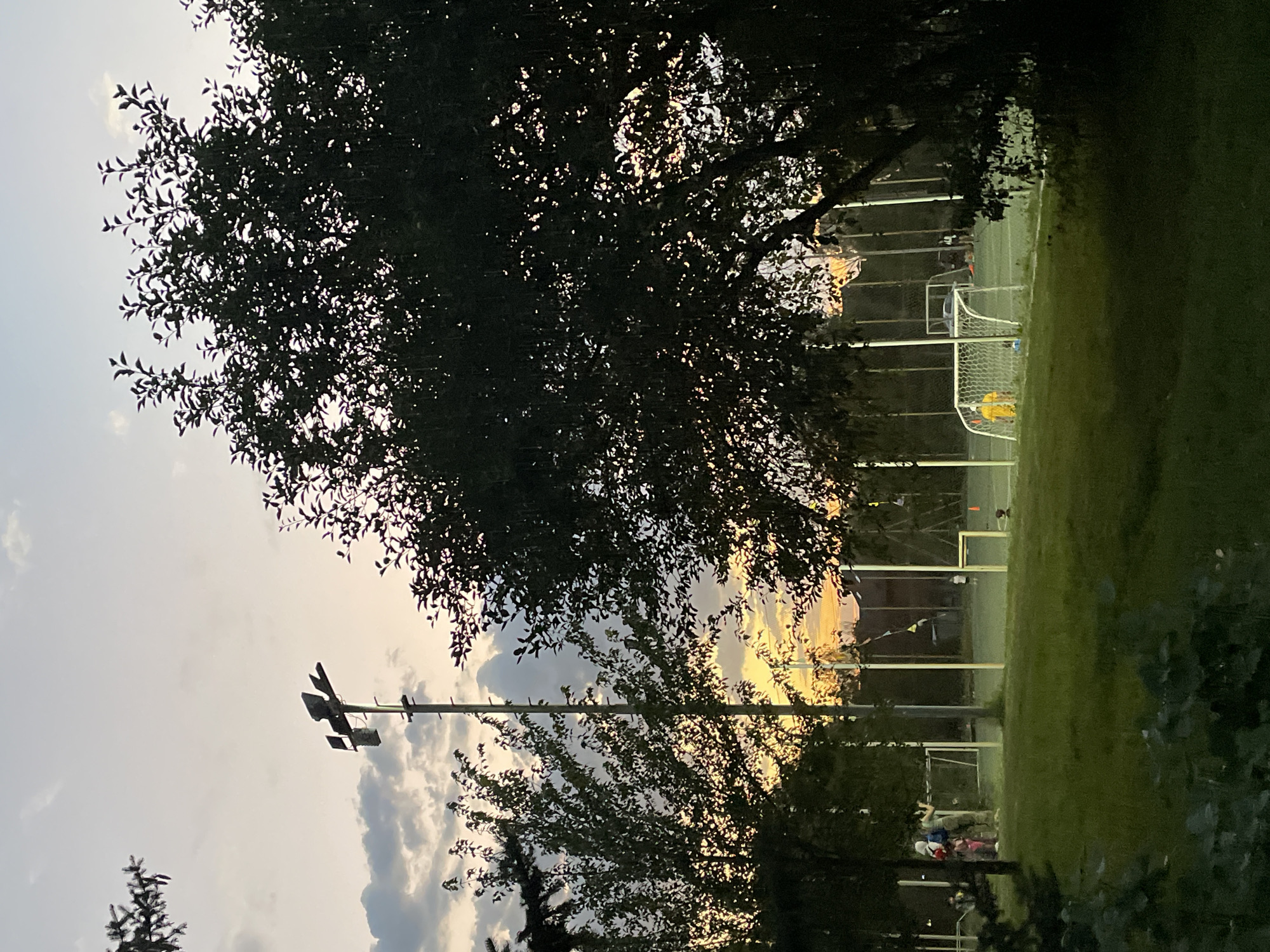 This image depicts a sports field, likely a soccer field, in Almaty, Kazakhstan, during dusk. The sky is painted with warm hues of orange and yellow from the setting sun, contrasted by patches of gray clouds. Tall floodlights stand sentinel over the field, illuminating sections of the green grass and the white boundaries. Soccer goals with white nets are visible, one with a person in a yellow shirt standing in front of it. Several individuals, some appearing to be children, are present on the field, engaged in activities that suggest a game or practice is underway. The foreground is dominated by lush green grass and the dark silhouettes of trees, adding a sense of depth and framing the scene. The overall atmosphere is serene, capturing a moment of recreational activity as daylight fades.