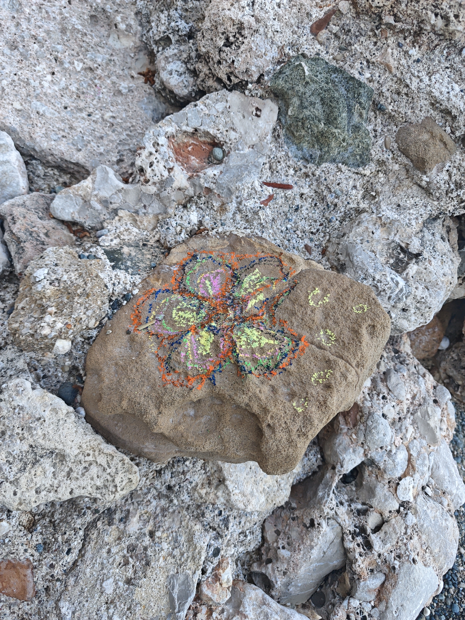 This image displays a close-up view of a large, brown, sandy-textured rock adorned with a vibrant, colorful drawing. The drawing resembles a stylized flower or mandala, executed in bright chalk or pastel colors including orange, pink, yellow, blue, and green, with a central pink and green section surrounded by layered, contrasting color outlines. To the right of this main drawing, several smaller, circular yellow marks are visible on the rock's surface.
The central decorated rock is nestled among a collection of various other rocks and debris. These surrounding elements are mostly light gray or white, with some pieces showing porous textures or flecks of reddish-brown and green, suggesting a mix of natural stone and possibly concrete rubble. The overall ground is rough and uneven, composed of small pebbles and larger broken rock fragments.
The scene appears to be set outdoors during daylight hours, illuminated by natural, bright light. The rugged texture of the rocks and the sandy appearance of the main drawn rock, combined with the given location context of Kemer, Turkey, suggest a coastal area, perhaps a rocky beach or shoreline where natural erosion or human activity has left various rock types. No people are visible, and the activity depicted is the act of drawing on a rock, leaving a piece of temporary, colorful art in the natural setting. There is no discernible text visible in the image.