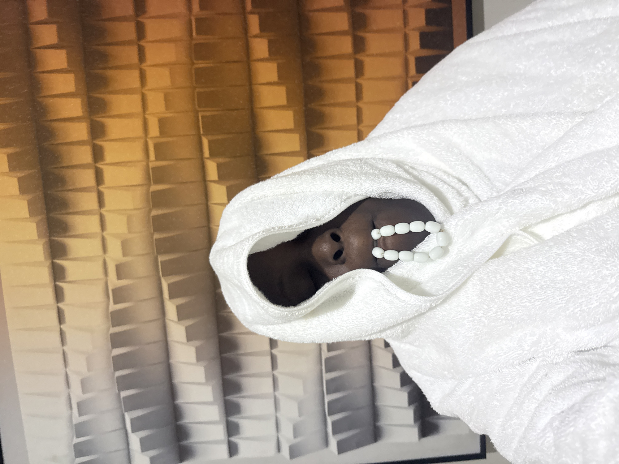 A person with dark skin is leaning back with their eyes closed, wearing a white hooded bathrobe and a necklace of white oval beads that rests on their lips. The background is a textured wall with a pattern of raised rectangular shapes, transitioning from light grey on the left to orange on the right.