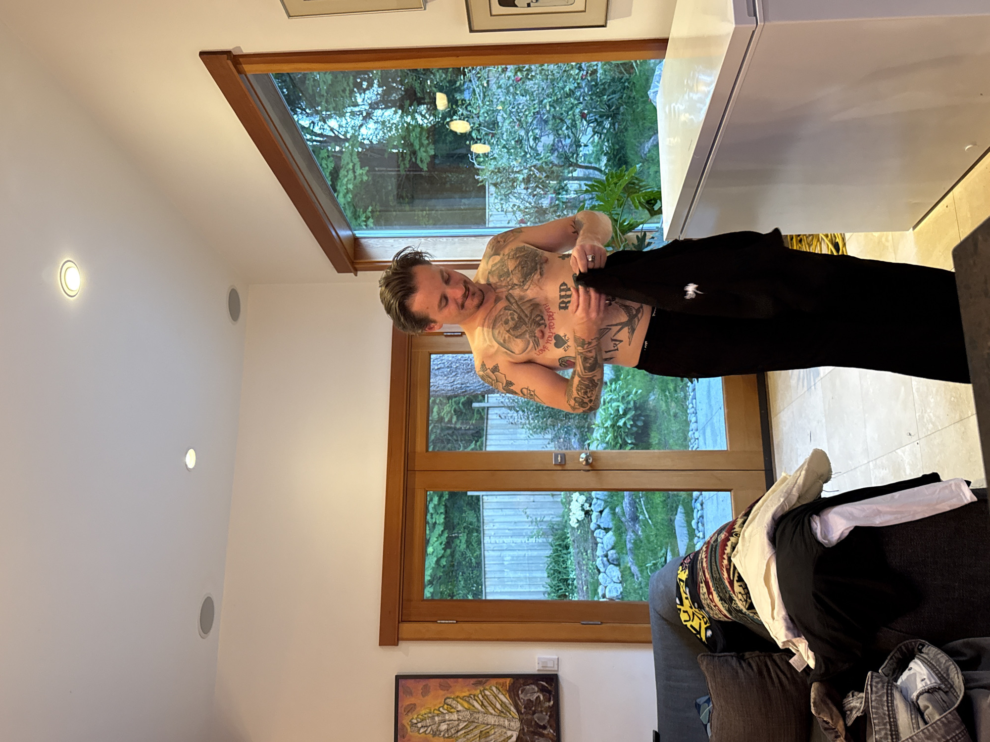 A person with tattoos is standing in a room, holding a black garment. The room features a large window offering a view of an outdoor garden. The location is suggested to be in the District of North Vancouver, Canada. The room also contains a couch covered with clothing and a painting on the wall.