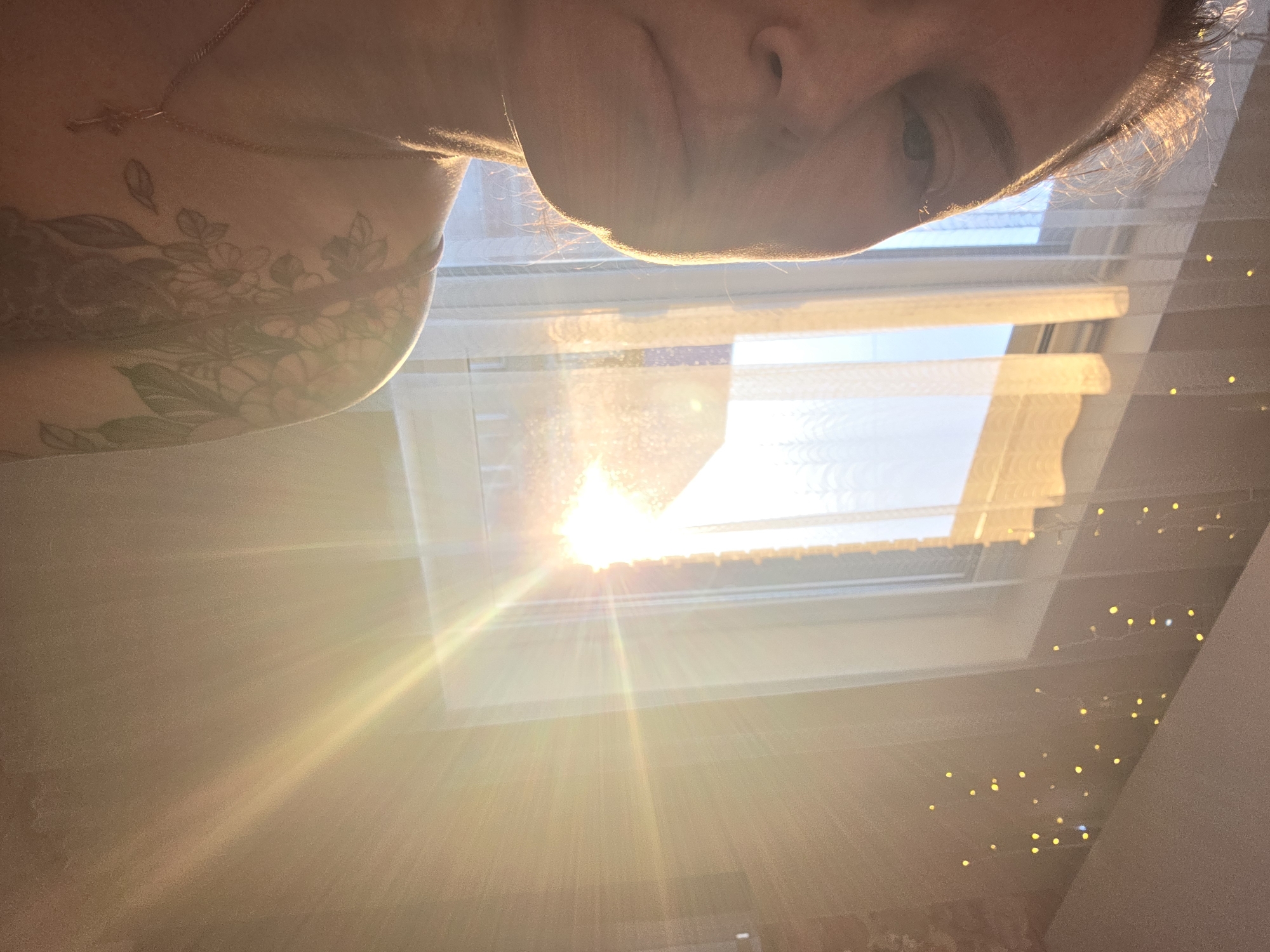 The image is a low-angle, close-up self-portrait. A person with fair skin is visible from the nose up, looking down at the camera. Parts of the neck and left shoulder are also visible, revealing a floral tattoo and a gold chain necklace with a small cross pendant. The background shows a bright, sunny window with white sheer curtains and blinds, suggesting it is daytime. The light streaming through the window creates a strong lens flare. A string of small, warm-toned lights is visible along a wall near the window. Without further information, it is impossible to confirm if the image was taken in the city of Chaikovsky, Russia.