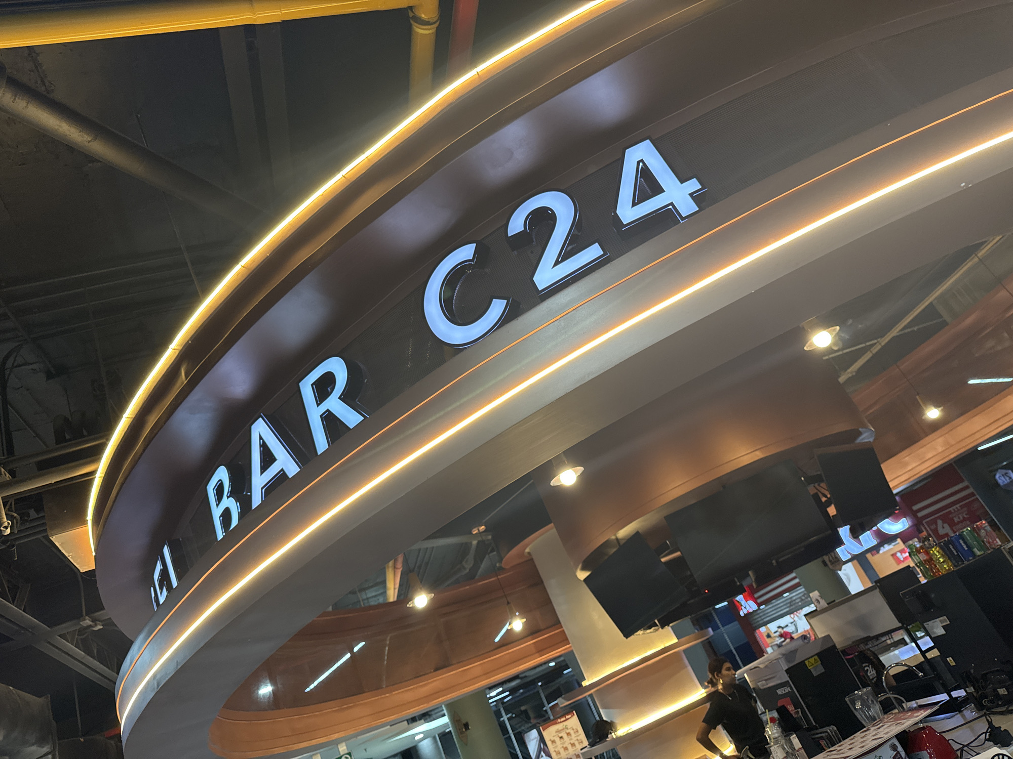 The image shows the "DEL BAR C24" sign, likely a bar or food establishment, located in what appears to be a shopping mall or public area. The sign is prominently displayed with white lettering against a brown background, illuminated by two light lines. Below the sign, there is a view into the bar area, with a person standing near a counter, possibly an employee. There is a display case behind the person, showing various items, and a red KFC sign is visible. The environment is well-lit and the angle of the photograph indicates a position looking up towards the sign from below. There is no specific information in the image that would indicate its location in Caracas, Venezuela.