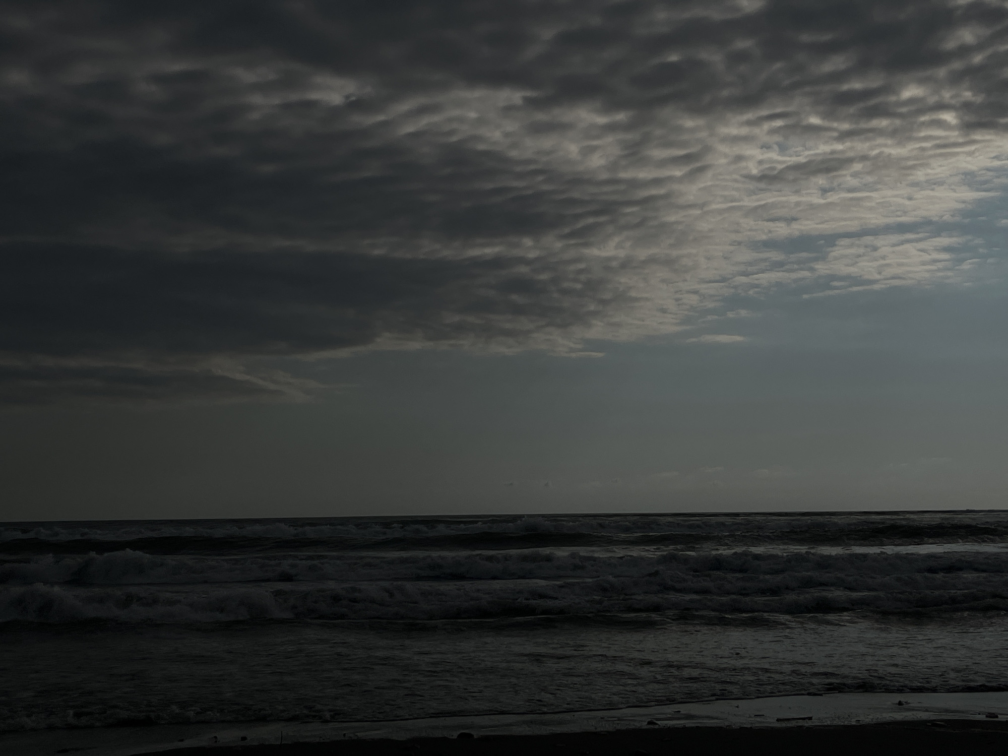 The image depicts a dimly lit, overcast seascape. Dark, textured clouds fill the upper portion of the frame, with a sliver of lighter sky visible towards the right. Below the sky, a series of dark, choppy waves roll towards a dark shoreline. The overall tone is somber and atmospheric, suggesting either dawn, dusk, or a storm-approaching weather condition. The location is identified as Sapsis Temi, Georgia.