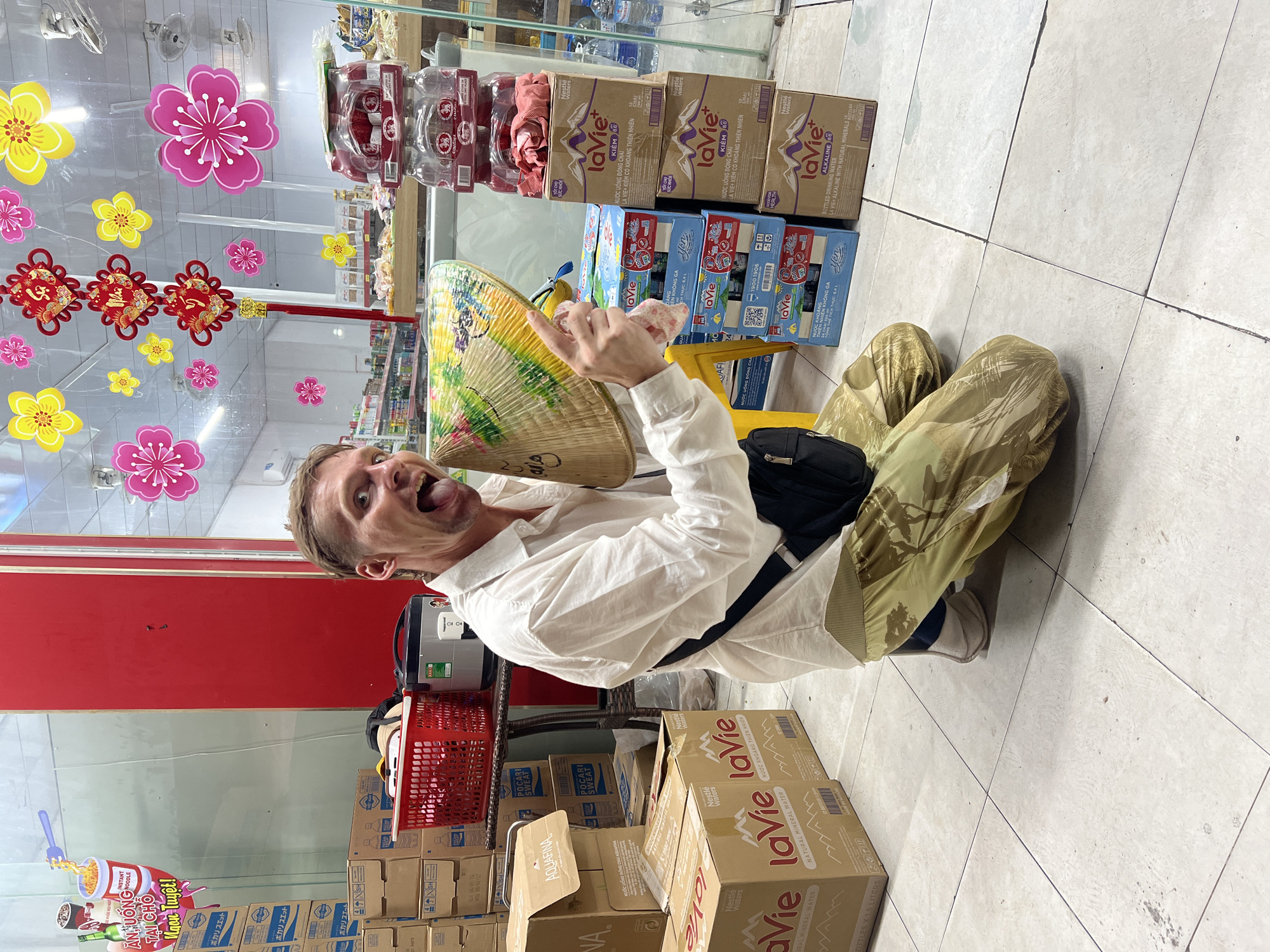 A man kneels on a tiled floor in front of a store display in Da Nang, Vietnam, holding a conical hat and sticking his tongue out at the camera. He is wearing a white button-down shirt and camouflage pants. The store appears to be selling bottled water and other beverages, with stacks of cases of La Vie water and other products visible. The store also has festive decorations, including pink and yellow paper flowers and red Chinese knot decorations, suggesting a celebration or holiday. A red plastic basket and a rice cooker are also visible near the man.