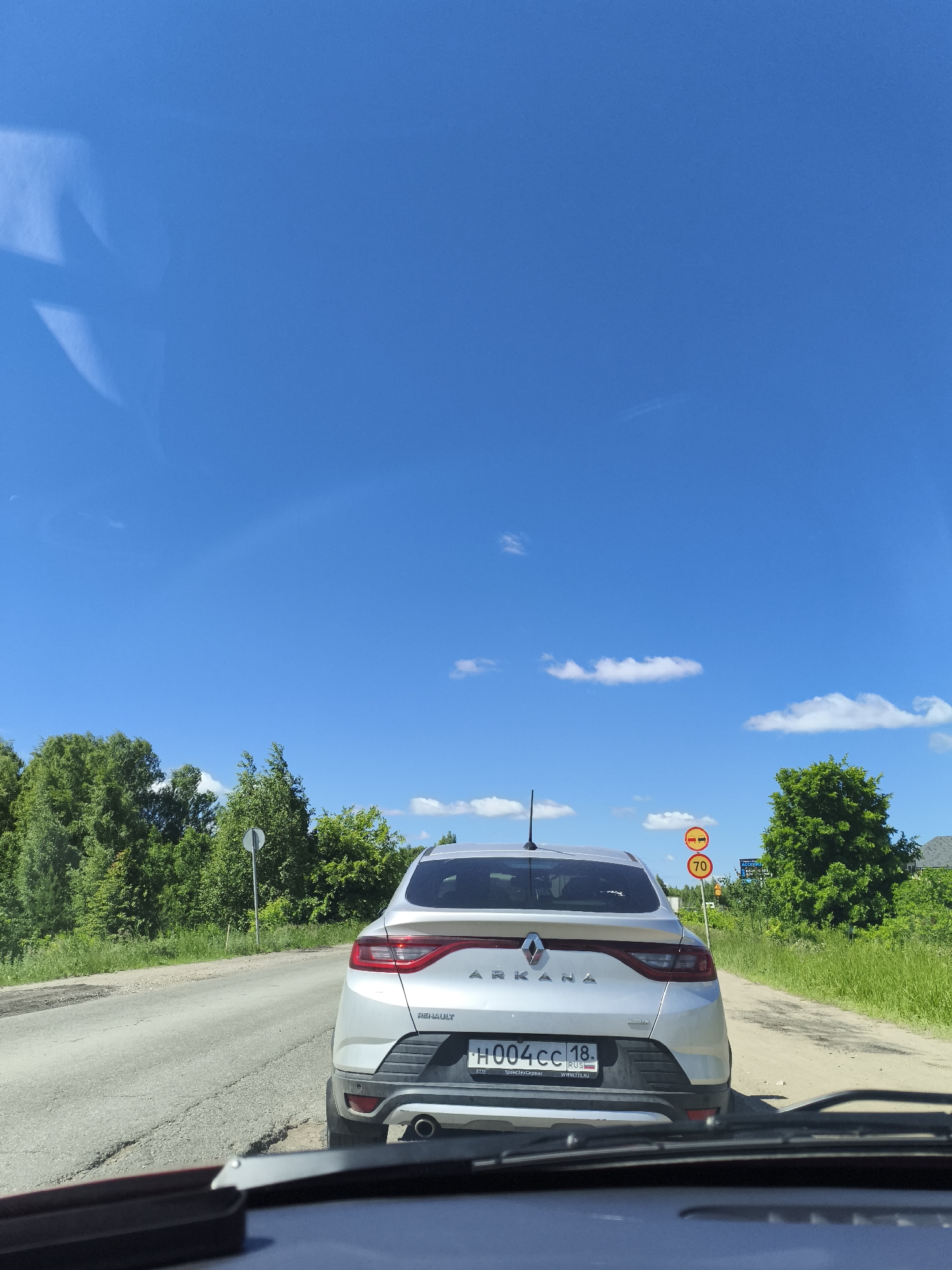 The image is taken from inside a vehicle, looking forward at a silver Renault Arkana car driving on a paved road in the city of Yagul, Russia. The car is in the foreground, slightly to the right of the center. The road is cracked and appears to be in disrepair. To the left and right of the road are lush green trees and vegetation, indicative of a rural or suburban environment. The sky is a clear, bright blue with a few scattered white clouds. There are two circular traffic signs visible on the right side of the road. The upper sign depicts two red circles with horizontal bars, signifying a restriction or prohibition, while the lower sign shows a red circle with the number "70" inside, indicating a speed limit of 70 kilometers per hour. The license plate of the Renault Arkana is partially visible, displaying Russian characters and numbers. The overall scene suggests a sunny day in a scenic, possibly remote, location.