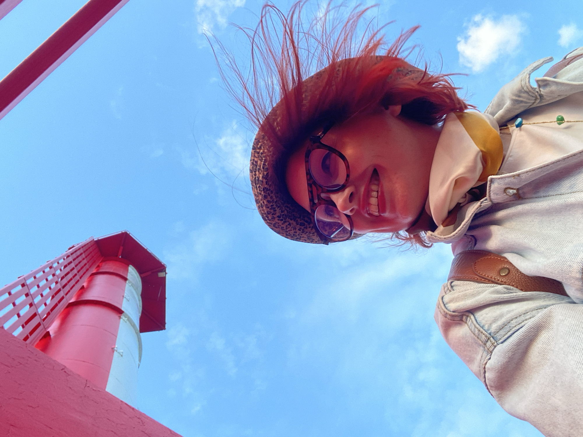 A low-angle shot shows a person and a lighthouse against a partly cloudy, blue sky. The person, possibly female, wears a hat, glasses, a jacket, and a scarf. Their red hair sticks up from under the hat. They are smiling. The red and white lighthouse is located in Sochi, Russia. A red railing runs along the bottom-left corner of the frame.