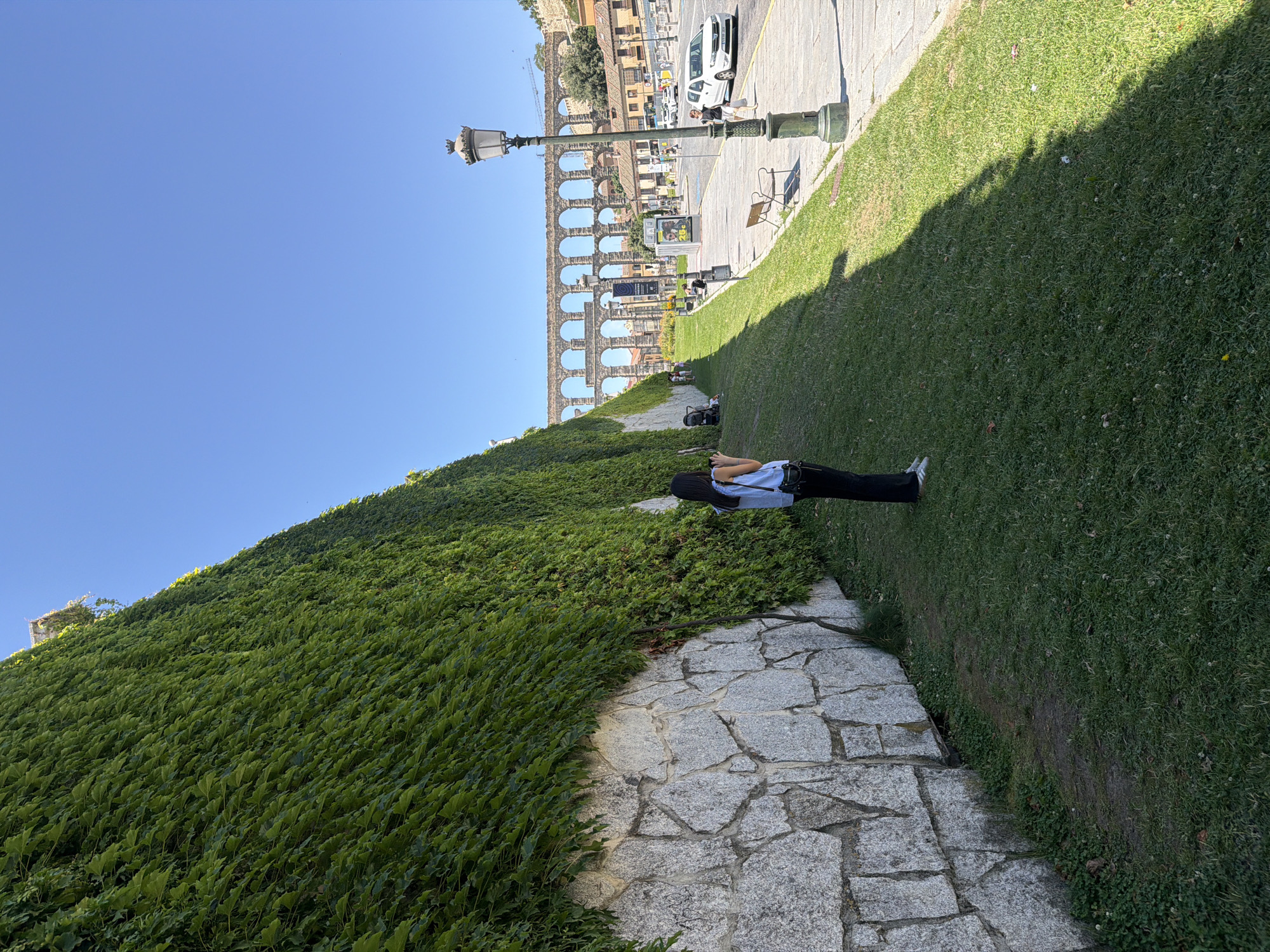 The image shows a woman standing on a grassy slope in front of a stone wall covered in green ivy. In the background, the iconic Aqueduct of Segovia is visible, spanning across the city under a clear blue sky. The aqueduct is a long, multi-tiered structure with numerous arches, characteristic of Roman engineering. To the right of the aqueduct, buildings and a street with cars and pedestrians can be seen. A lamppost with an ornate top stands in the foreground on the right side of the image. The scene appears to be a sunny day in the historic city of Segovia, Spain.
