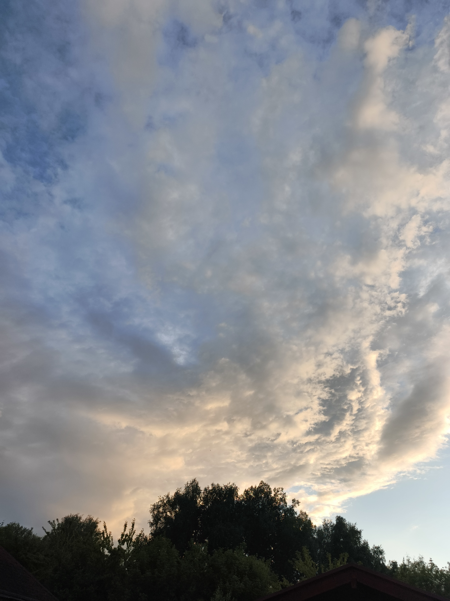 The image depicts a sky filled with a mix of light and dark clouds, with patches of blue visible. The clouds are illuminated by a soft, warm light, suggesting either sunrise or sunset. In the lower portion of the image, the dark silhouettes of trees and a roofline are visible against the lighter sky. The scene appears to be an outdoor view, possibly from a residential area, in Катунь, Russia. No people or specific actions are identifiable.