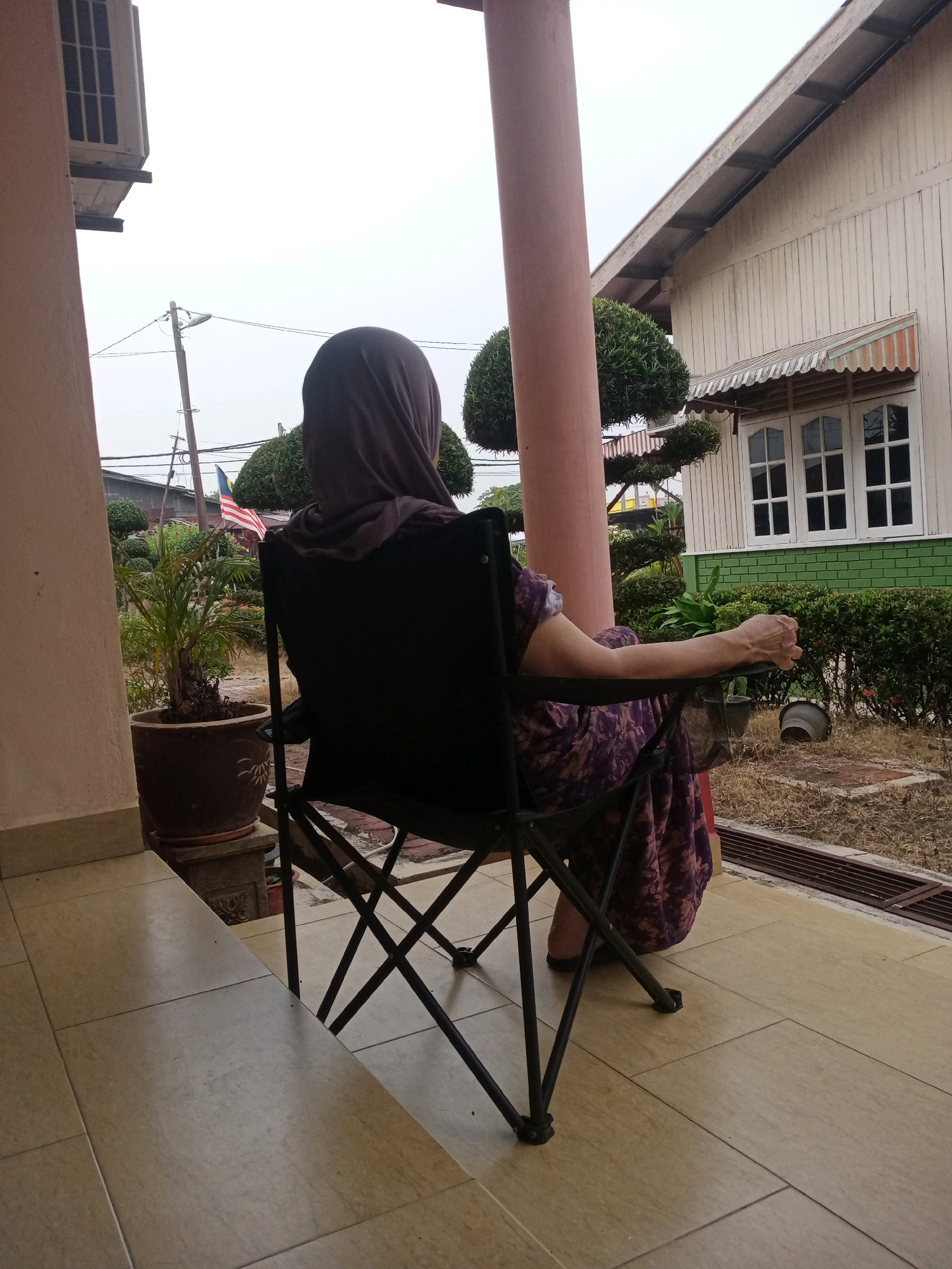 A person wearing a hijab and a purple patterned dress is sitting in a black folding chair on a tiled porch. The person is facing away from the camera, looking towards a house and a garden. The house is white with green trim and has several windows. There are manicured bushes and trees in the garden, as well as a potted plant. In the background, a Malaysian flag is visible on a flagpole. The sky is overcast. The scene appears to be in a residential area in Kampung Guar Batu Hitam, Malaysia.