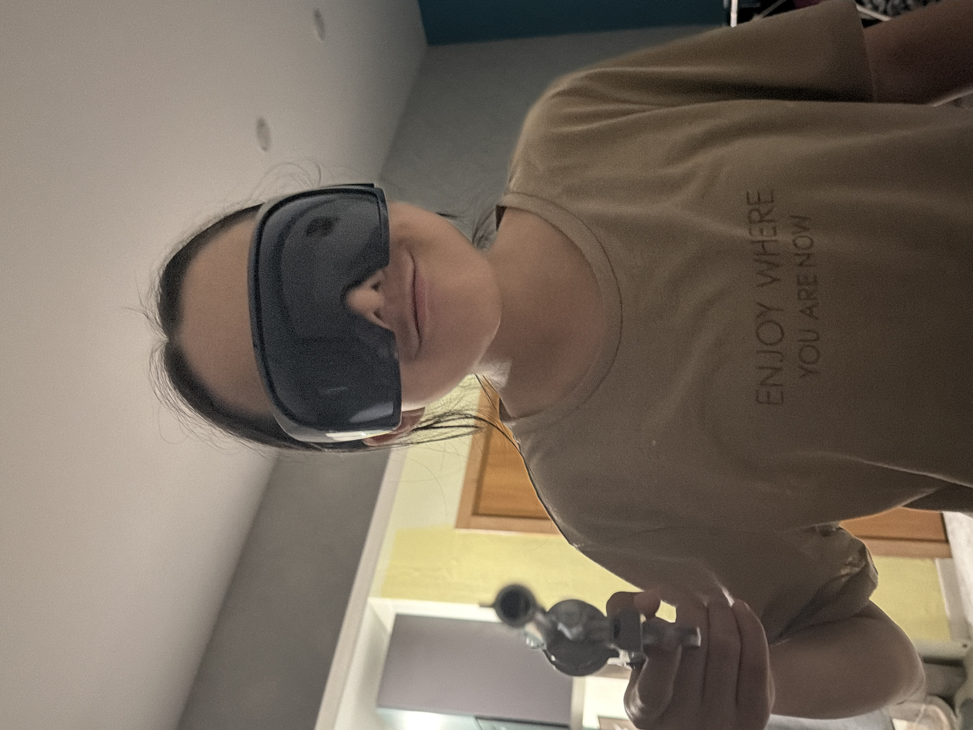 A person wearing black eye coverings is holding an object that appears to be a glass pipe in their right hand. The person is wearing a beige T-shirt with the words "ENJOY WHERE YOU ARE NOW" printed vertically on the chest. The background includes a wall, a door frame, and what appears to be a kitchen area. The location in the city is unknown.