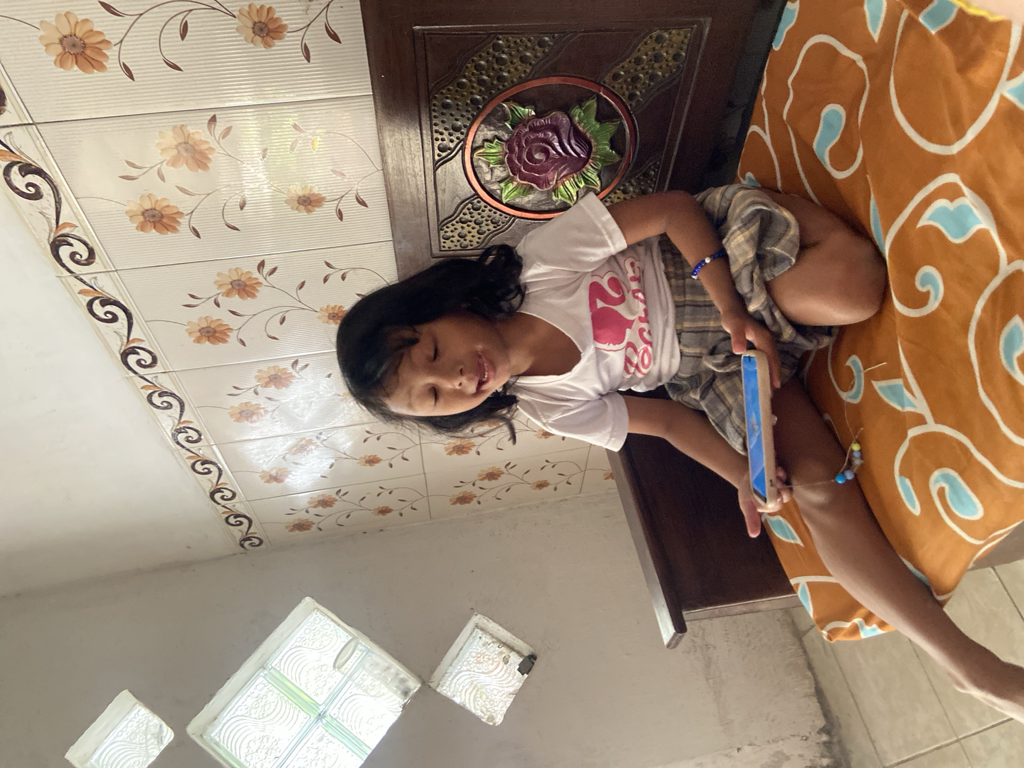 In the city of Baron, Indonesia, a young girl with dark hair sits on a patterned orange cushion. She is wearing a white t-shirt with a pink graphic and a plaid skirt. Her eyes are closed, and her mouth is open as if she is speaking or singing. She holds a tablet computer in her hands, which are resting on her lap. The girl is positioned in front of a wooden bench with a decorative carved rose motif. The background features a wall tiled with a floral pattern and two small windows with textured glass. The overall scene suggests a casual, indoor setting.