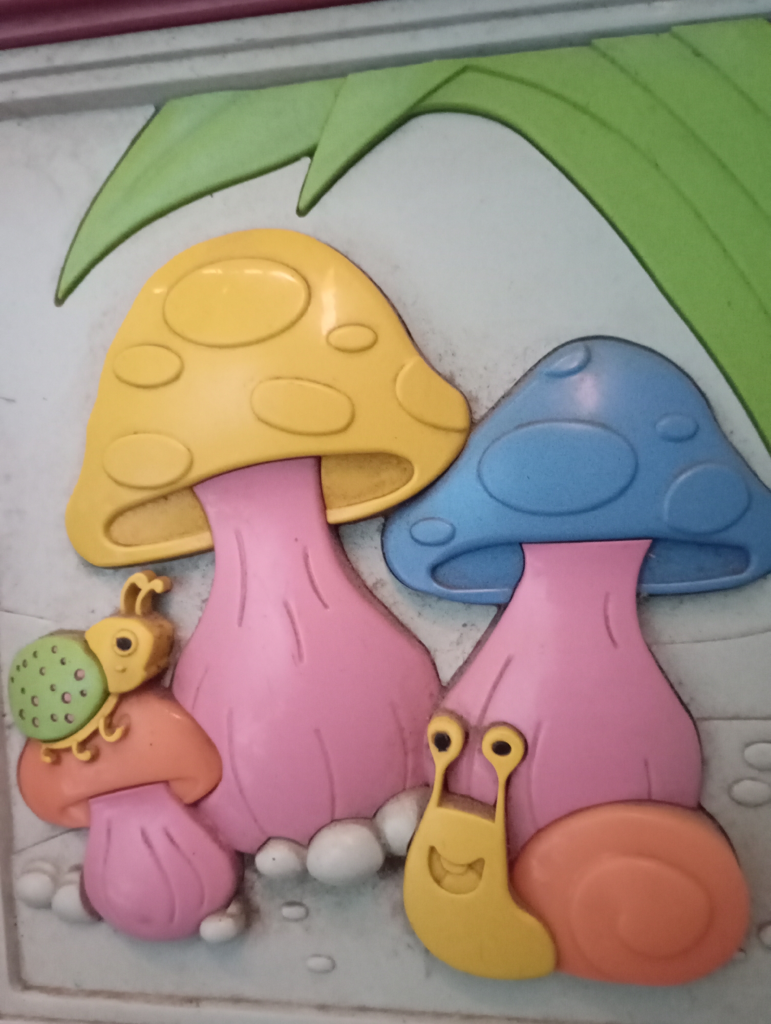 The image is a close-up, overhead shot of a decorative relief, likely a wall plaque or part of a playground structure, featuring colorful, cartoonish characters and elements. The main subjects are two mushrooms with pink stems and different colored caps: one yellow with oval indentations, and one blue with oval and circular indentations. To the left of the yellow mushroom, a small, green ladybug-like character with yellow antennae and eyes is perched on a small orange and pink mushroom. To the right of the blue mushroom, a yellow snail with large black eyes and a smiling mouth is positioned next to its orange, spiraled shell. Several small white oval shapes resembling pebbles or dew drops are scattered around the base of the mushrooms. In the upper portion of the image, stylized green leaf shapes are visible against a light gray background. The overall scene depicts a whimsical, natural setting with animated creatures. There is no indication or context related to Kecamatan Haurgeulis, Indonesia in the provided image.