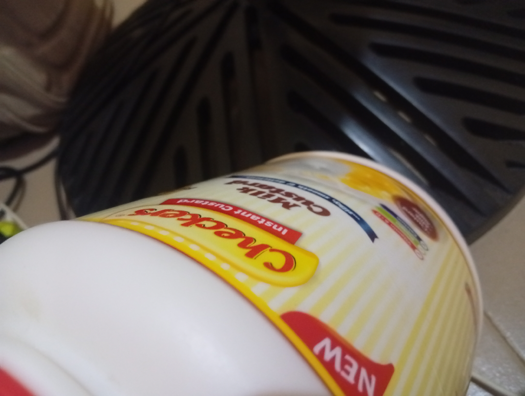 In Ukukwa, Nigeria, a white plastic container of "Checkers Instant Custard" is positioned at an upward angle, with its yellow and white striped label prominently displayed. The brand name "Checkers" is written in red lettering on a yellow oval. The background consists of a blurred, dark, ribbed object, possibly a grill or a textured surface. The lighting is dim, casting shadows and highlighting the reflective surface of the container.