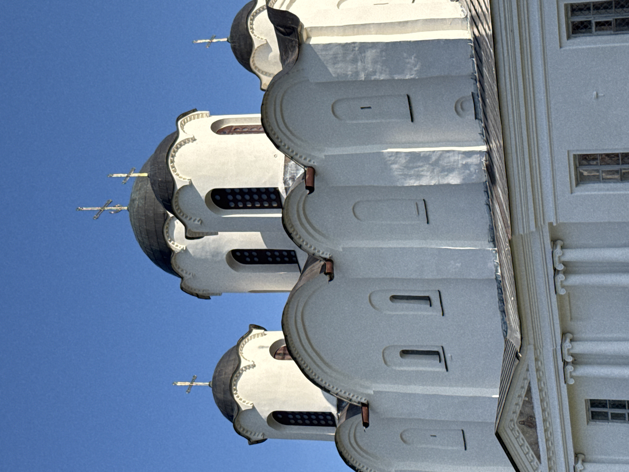 The image shows a close-up, angled view of the white exterior of the St. Sophia Cathedral in Veliky Novgorod, Russia. The cathedral features multiple domes, with the central and largest dome being dark and topped with a cross. Smaller white towers with dark domes also rise from the structure, each also bearing a cross. The architectural style is characteristic of Russian Orthodox churches, with arched windows and decorative elements along the rooflines. The sky is a clear, bright blue. The lower portion of the image reveals more of the building's facade, including classical columns and arched windows. The overall scene is one of architectural grandeur under a clear sky.