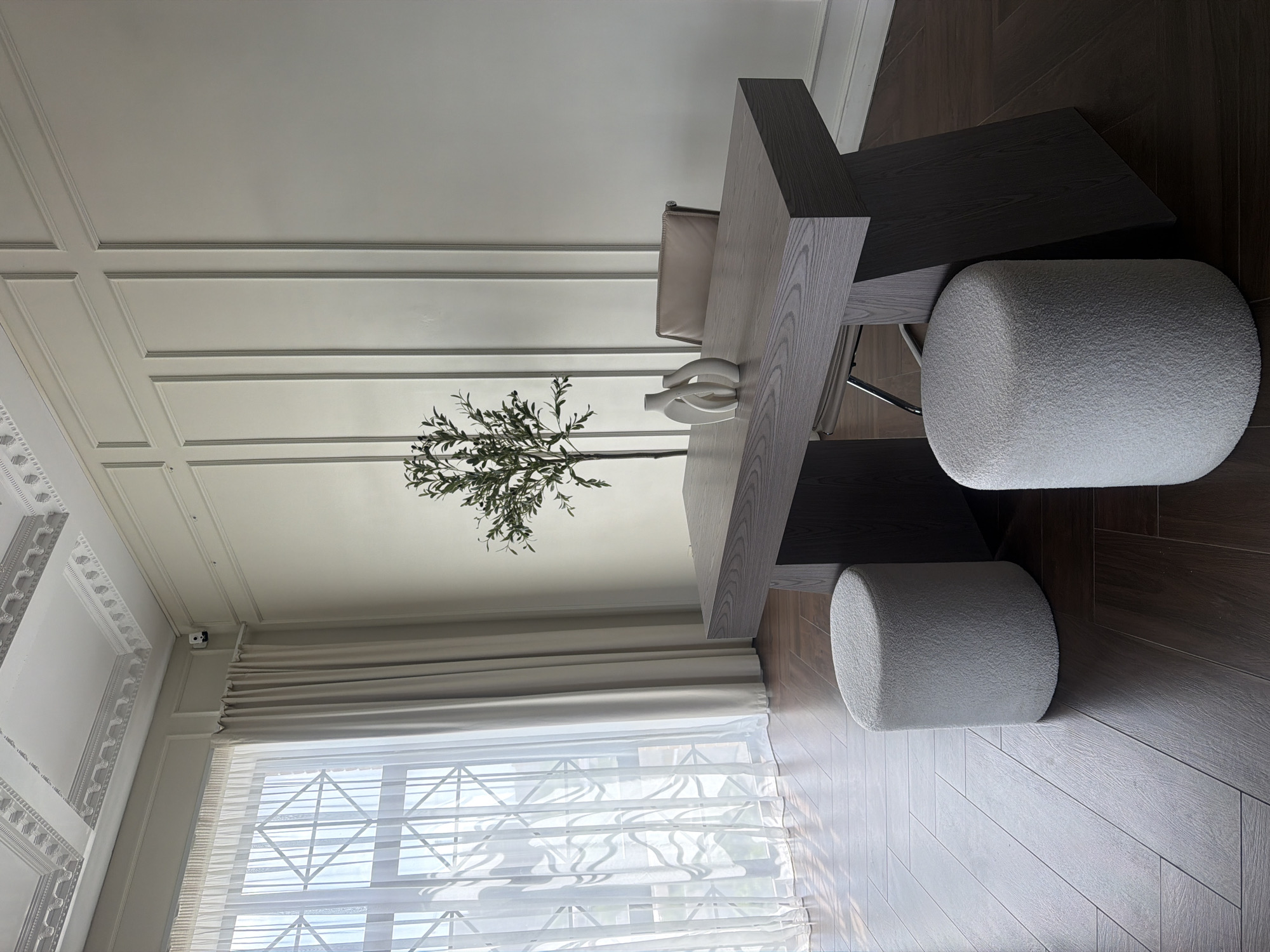 A rotated indoor view captures a scene with a modern aesthetic. The image features a geometrically shaped table with a light grey tabletop and dark grey support structure. On the table are decorative vases and a plant. Two white, cylindrical, textured ottomans are positioned around the table. A chair with a light-colored seat is partially visible behind the table. The room is well-lit by natural light coming through a large window covered by a light-filtering Roman shade. The floor is a dark wood. The wall has wainscoting. A security camera is mounted near the window. The location is likely indoors, possibly Rostov-on-Don, Russia.