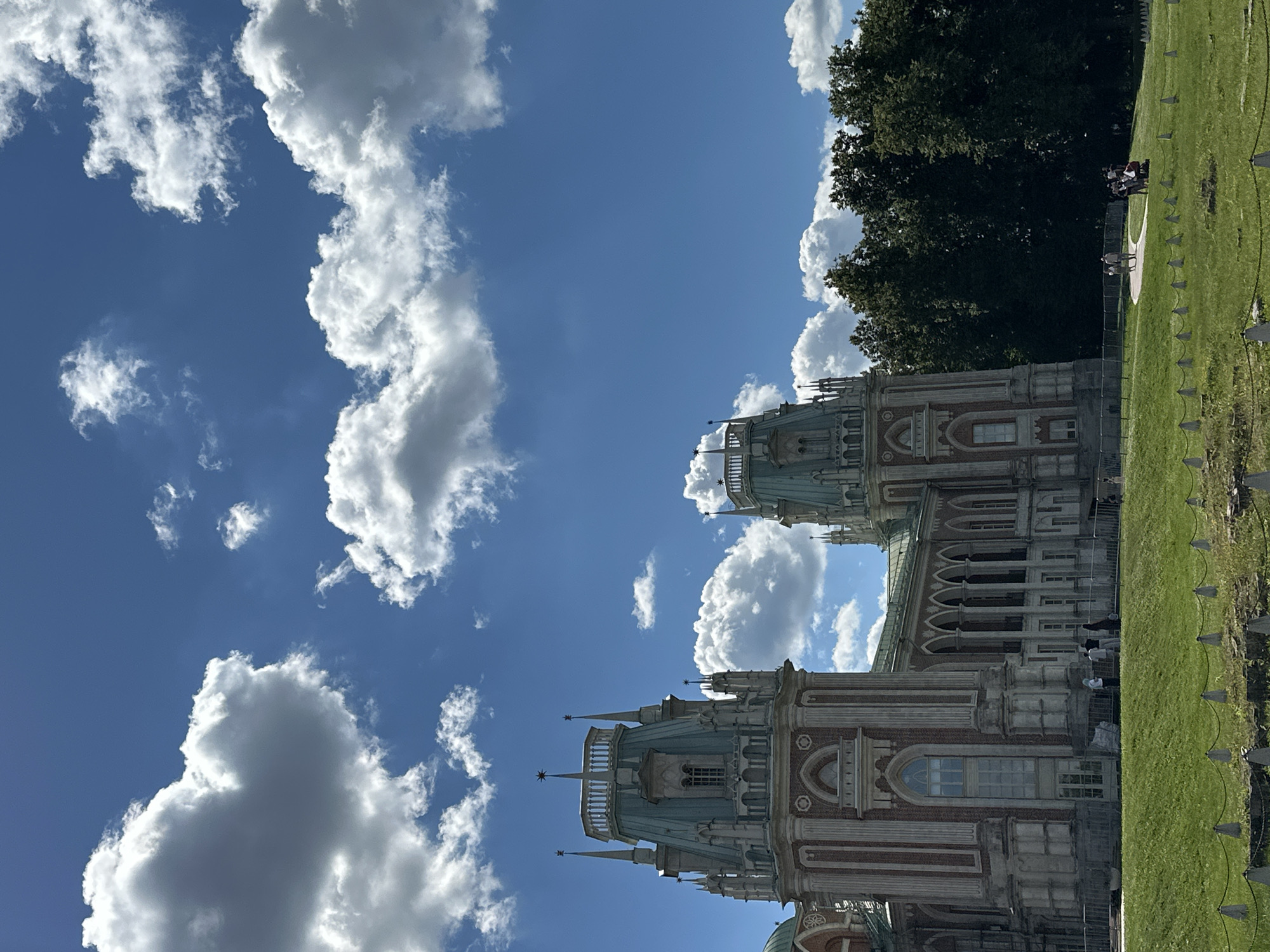 The image shows a scenic view of a grand architectural complex with a lush green lawn and a vibrant blue sky dotted with fluffy white clouds. The architecture appears to be of historical significance, with intricate details, towers, and arches, hinting at a palace or a museum. The main subjects are the buildings and the sky. There are a few people visible in the background, scattered on the lawn and near the building, seemingly enjoying the outdoors. Some are walking, and a small group appears to be gathered in the distance.
The time of day is clearly daytime, suggested by the bright sunlight casting shadows and illuminating the scene. The weather seems pleasant, with a clear blue sky and scattered clouds, indicative of a fair-weather day. The presence of green grass and the attire of the people suggest a warm season, likely spring or summer.
Notable details include the distinctive red brick and white stone facade of the buildings, adorned with ornate architectural elements like columns, arched windows, and spires. The extensive lawn area suggests a park or garden setting, common around historical estates. The fence with small triangular markers along the edge of the lawn creates a boundary. The dark green foliage of a large tree on the right side of the image provides a contrast to the lighter tones of the buildings and the sky.
The overall mood conveyed by the image is one of tranquility and beauty, highlighting the architectural grandeur and the pleasant natural surroundings. No visible text is present in the image. Based on the provided location context, this scene is likely in Moscow, Russia, possibly a historical park or estate known for its architecture.