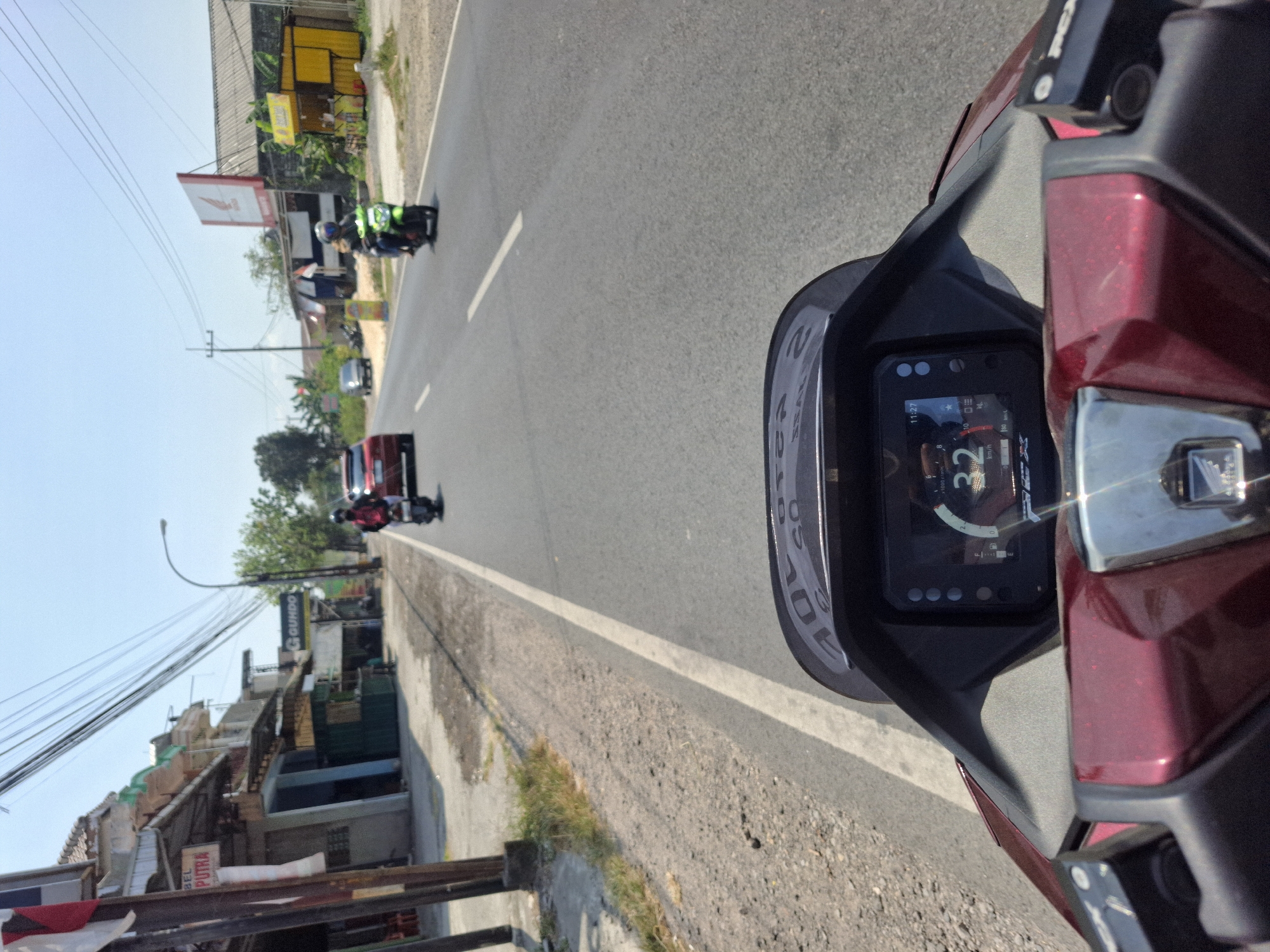 The image is a first-person view from a motorcycle in Banjarmadu, Indonesia. The motorcycle's dashboard displays a speed of 32 km/h and the time is 11:27. The rider is on a road with shops lining the left side and more shops and trees on the right. A red car is visible ahead, and two other motorcycles are on the road, one in front and one to the right. Power lines crisscross the sky.
