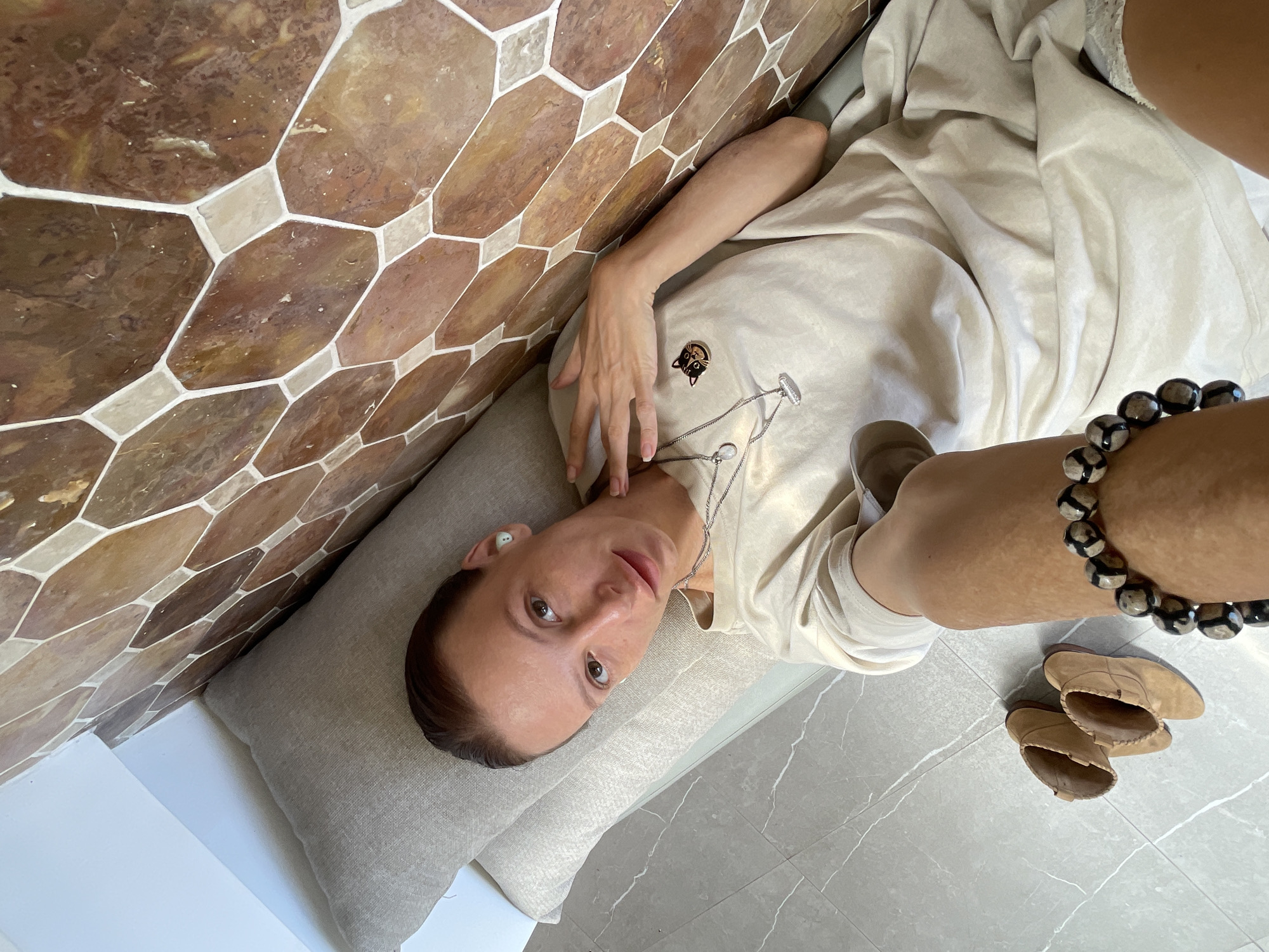 A woman lies on a grey cushion, adjacent to a decorative wall of brown and beige geometric tiles, and grey marble flooring. The woman wears a light-colored t-shirt with a cat logo, a silver necklace, and a white earbud in her left ear. She has brown hair pulled back, brown eyes, and pale skin. She holds her right hand up, fingers extended, toward her face. A chunky bead bracelet is visible on her right wrist. A pair of light tan leather shoes lie on the marble floor. The photograph appears to be taken in a residence in Ubud District, Indonesia.