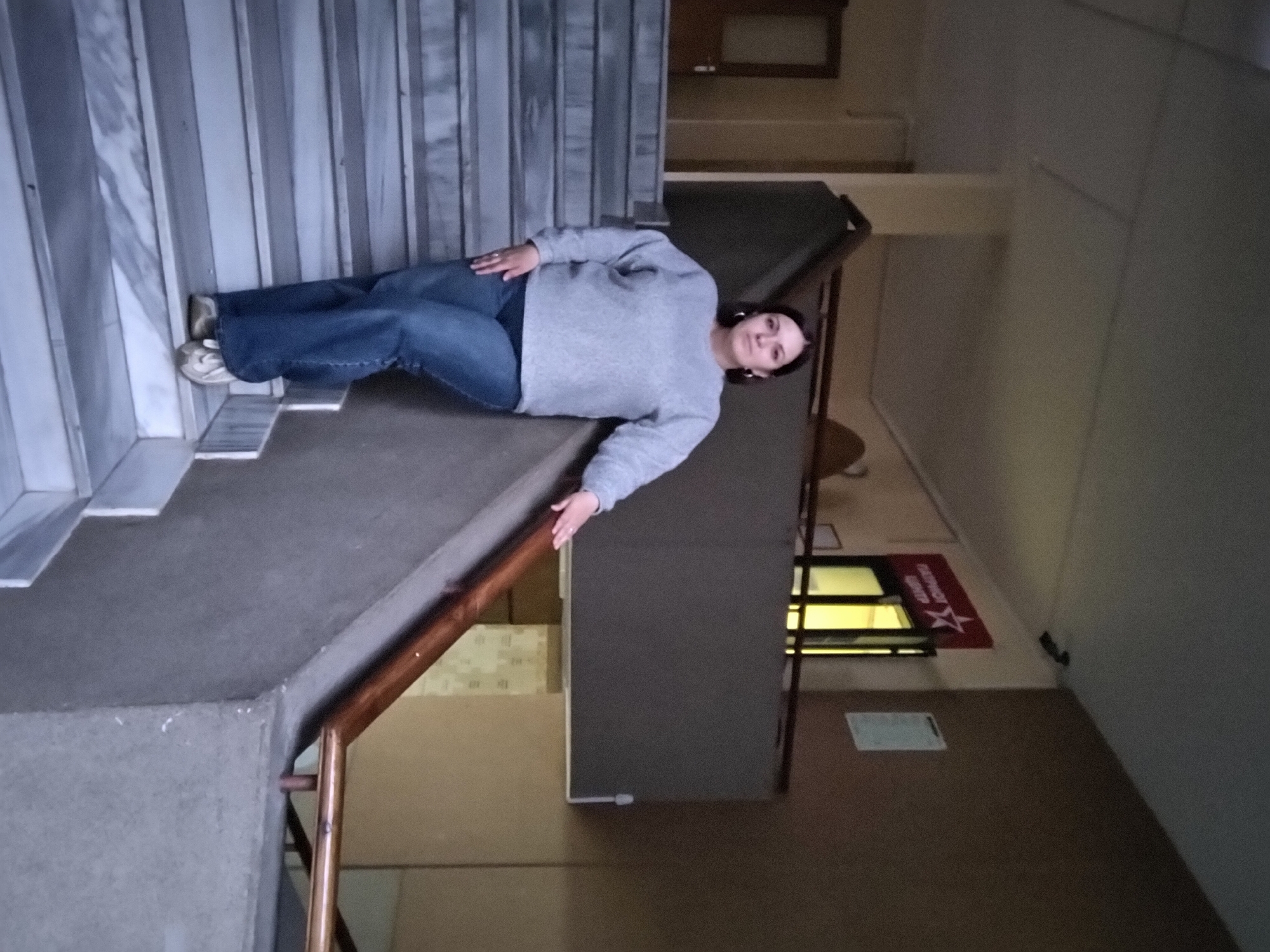 The image captures a woman lying on a staircase inside a building, likely in Markovskii, Russia. The woman, with dark hair, is wearing a light gray long-sleeved top, blue jeans, and white sneakers. She has a ring on her left ring finger. She is positioned with her head towards the right side of the frame and her feet to the left, looking directly at the camera with a calm expression. Her left arm is extended along the stairs, while her right arm rests across her body.
The staircase consists of grey, possibly marble or stone, steps on the left, transitioning to a darker, carpeted or concrete section on the right where the woman is. A wooden handrail runs along the right edge of these darker stairs. In the background, a textured wall is visible to the left of the woman. Beyond the stairs, there's a corridor or room with light-colored walls, and a dark partition or structure stands on the right. A window or opening in the distance emits a yellow light.
A key detail is a red mat or sign on the floor in the background, which displays Cyrillic text. The text reads "ПАТРИОТИЧЕСКИЙ ЦЕНТР" (Patriotic Center) and includes a star symbol, reinforcing the location context as Russia. The scene is indoors, with artificial lighting, suggesting a casual, posed photograph within a public or institutional building.