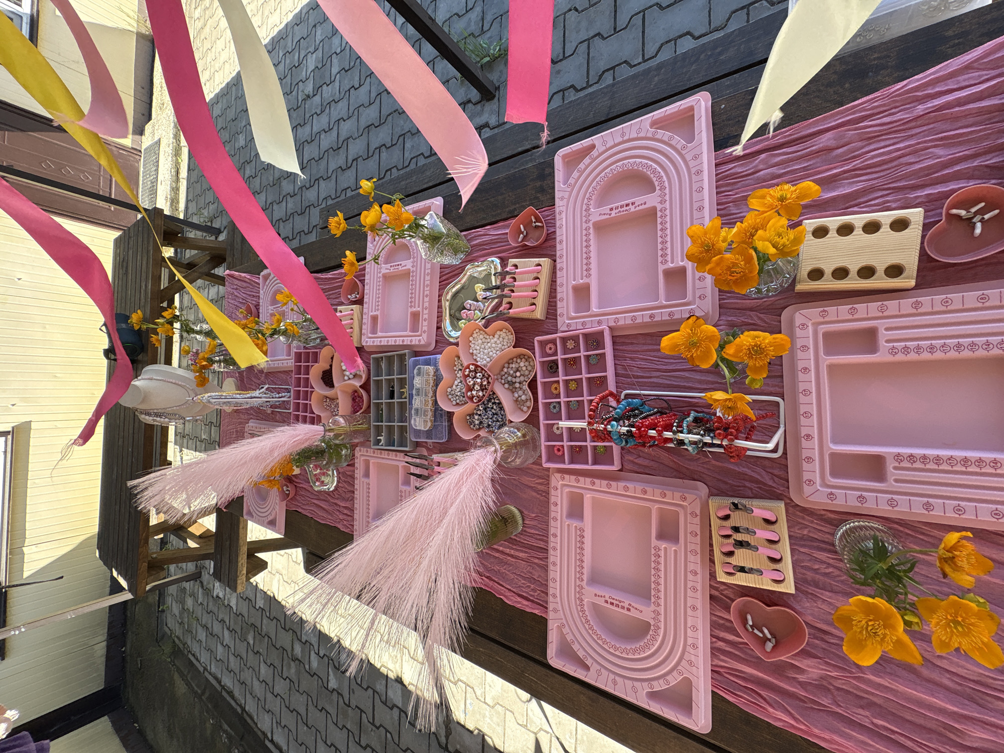 The image depicts an outdoor setting, likely a patio or garden, with a wooden picnic table covered in a pink tablecloth. The table is laden with various items for jewelry making, including pink plastic design boards, trays filled with beads of different colors and sizes, small tools like pliers and cutters, and several small vases containing orange flowers. Pink, yellow, and white ribbons are draped across the scene, suggesting a festive or celebratory atmosphere. In the background, a yellow building with a dark door is visible. The ground is paved with interlocking gray bricks. The overall impression is that of a jewelry-making party or workshop setup. No specific location in Birobidzhan, Russia is discernible from the visual cues.