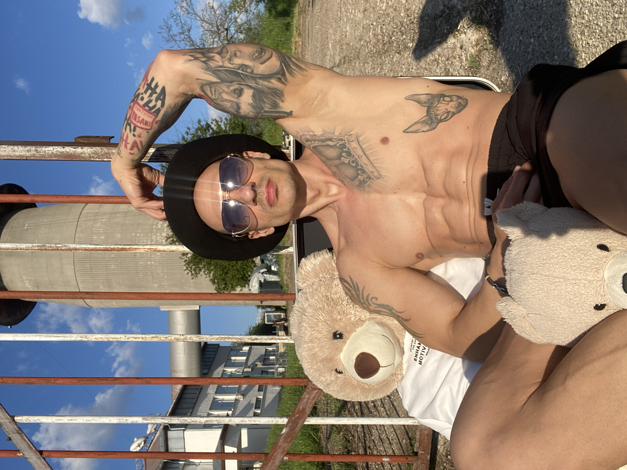 The image shows a shirtless man with multiple tattoos, wearing a black hat and sunglasses. He is sitting on a chair, holding a teddy bear. The background includes a metal fence, a large cylindrical structure, a building, and a blue sky with clouds. The location appears to be outdoors, possibly in an urban area. While there is not enough information to confirm the location as Pleven, Bulgaria, there are clues suggesting that the building behind the man is similar to buildings there, such as apartment buildings and industrial structures. The image lacks specific identifiers to confirm its precise location within Pleven.