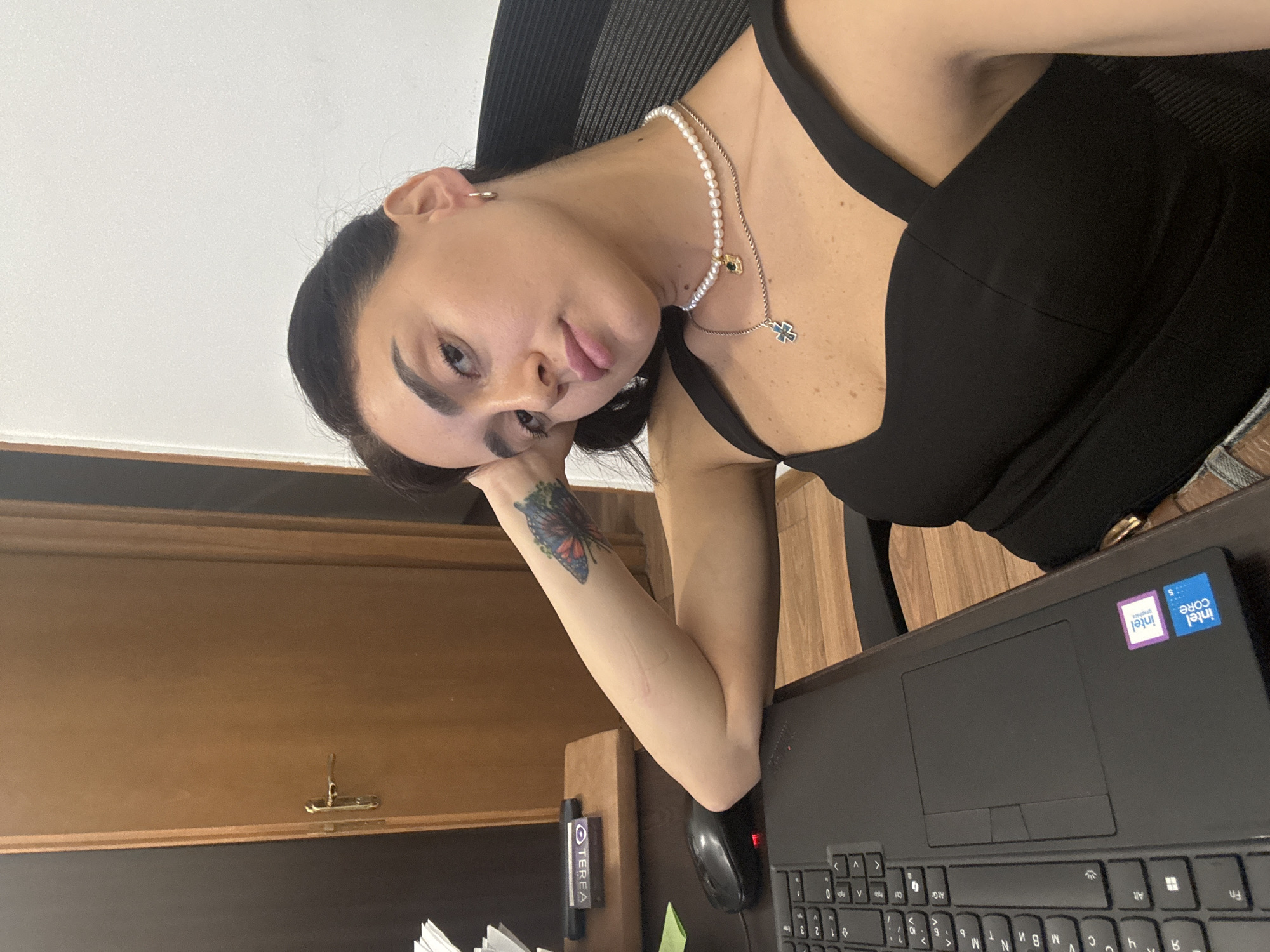 A young woman with dark hair and a butterfly tattoo on her left arm is sitting at a desk with a laptop, mouse, and a box of TEREA. She is wearing a black top and a pearl necklace with a cross pendant. The desk is in a room with a wooden door and white walls. The scene appears to be an office or home office. There is no visible indication of the specific location within the city of Tbilisi, Georgia.