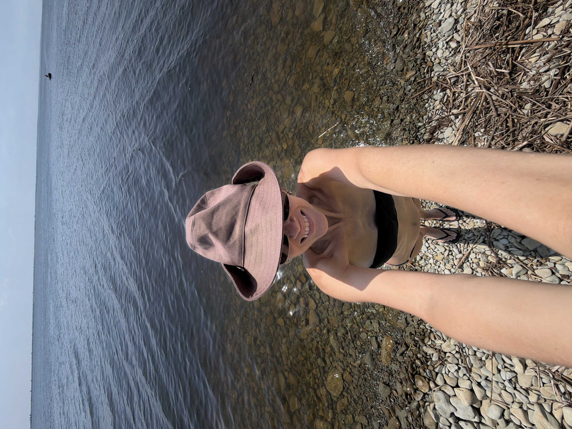 The image shows a person taking a selfie on a pebble beach next to a body of water. The person is wearing a wide-brimmed hat and sunglasses, and is smiling at the camera. They are also wearing a black bikini top. The water appears to be calm, with ripples on the surface. In the distance, a small boat is visible on the water. The beach is covered in smooth, grey and brown pebbles, and there are some dry branches and sticks scattered on the ground. The sky is clear and blue. The scene is likely a lake or a wide river, given the absence of visible waves and the distant horizon. The location is likely the Volga River, which flows through the city of Kamyshin, Russia.