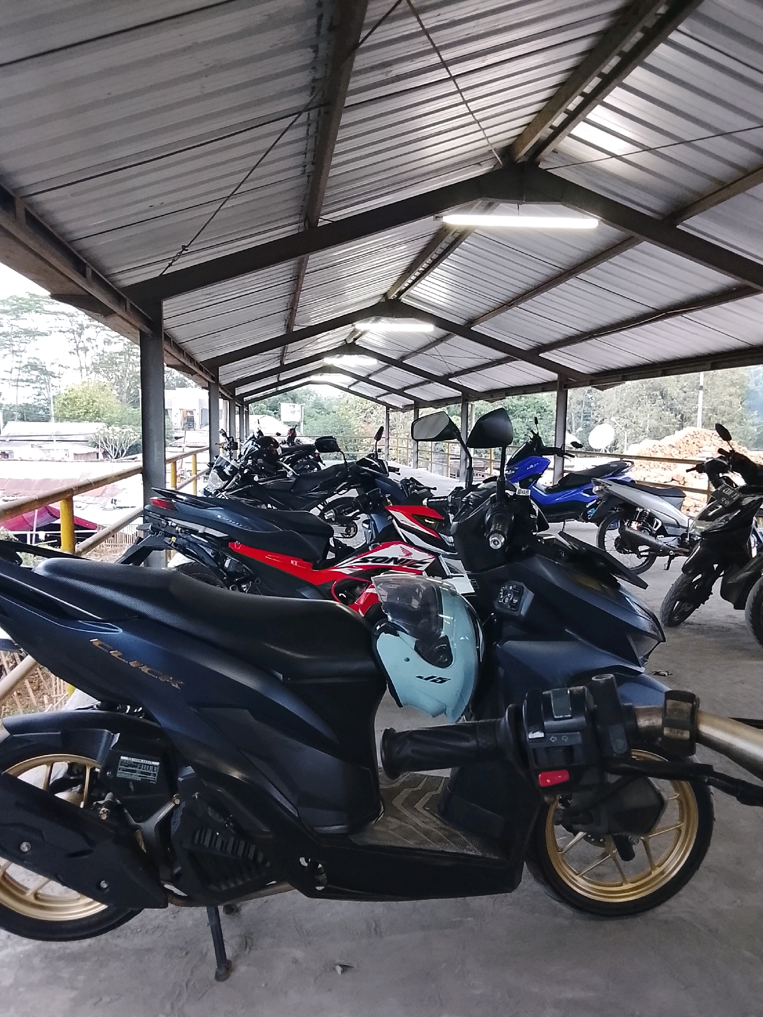 The image depicts a covered outdoor parking area, likely in Banjar, Indonesia, filled with numerous motorcycles. The structure is supported by metal beams and has a corrugated metal roof, with fluorescent lights visible overhead. In the foreground, a dark-colored scooter is parked, featuring gold rims on its wheels and a light blue helmet on its handlebars. Behind it, other motorcycles, including a red and white one, are lined up. The background shows trees, foliage, and some buildings, suggesting an urban or semi-urban environment. The lighting suggests it is daytime, possibly late afternoon given the slight dimness.
