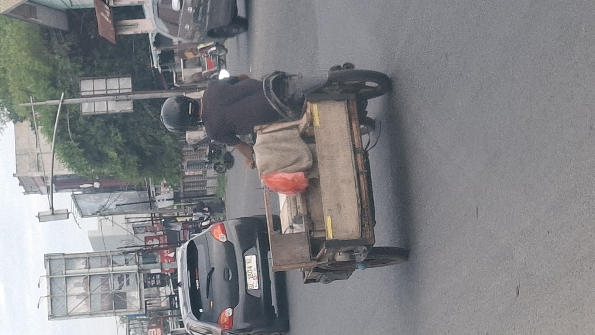 A man wearing a black helmet and a dark t-shirt rides a motorcycle with a trailer in Lhokseumawe, Indonesia. The trailer is made of wood and is carrying various items, including a red plastic bag and some fabric. The motorcycle is on a paved road, with a dark-colored hatchback car visible in the background. Buildings, billboards, and trees line the street, suggesting an urban environment. The sky is overcast, indicating possibly cloudy weather.