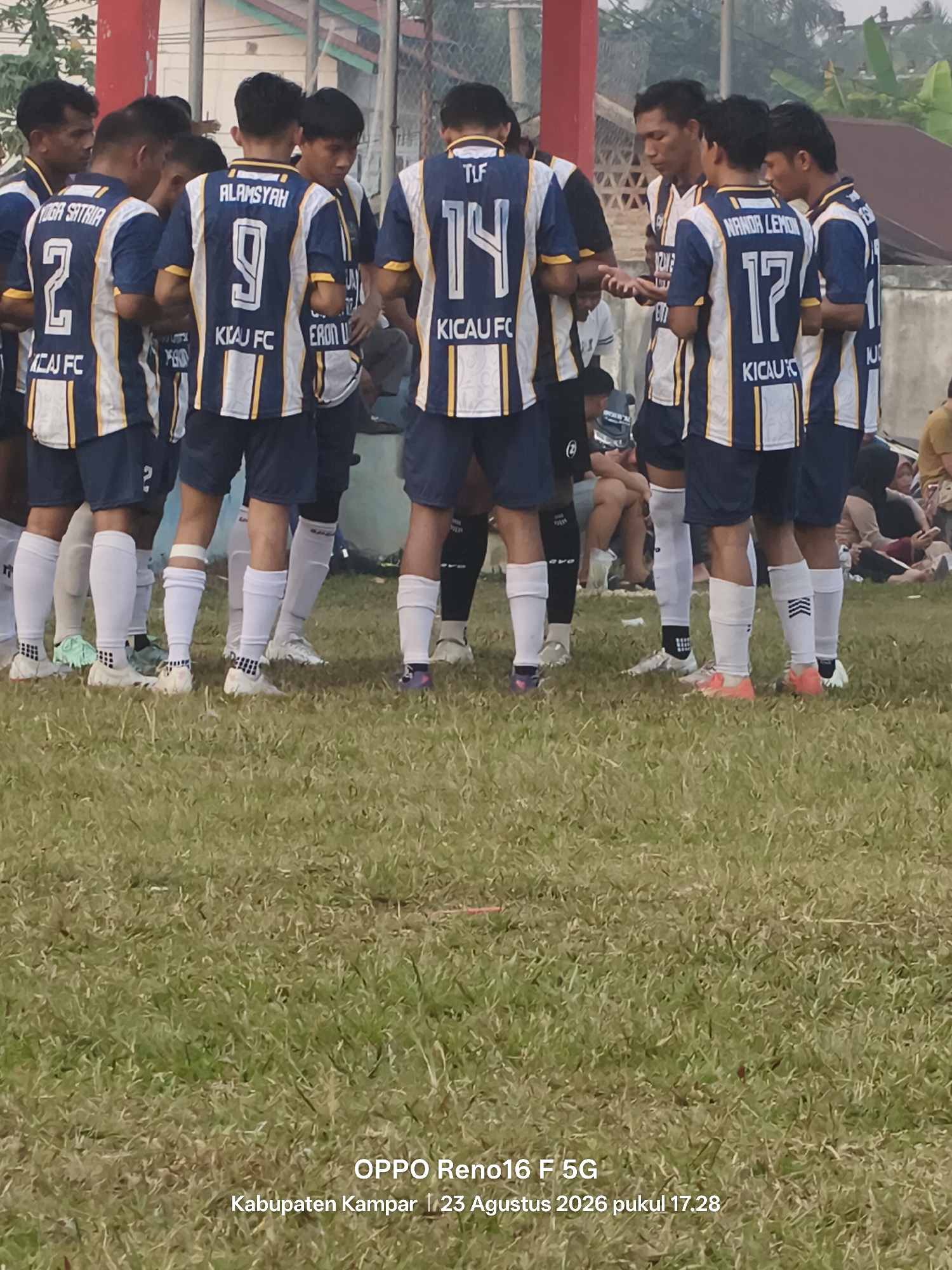 In Simalinyang, Indonesia, a group of young men in matching blue and white striped soccer uniforms are gathered in a huddle on a grassy field. The uniforms feature prominent numbers and names on the back, including "2 YOGA SATRIA," "9 ALAMSYAH," "14 TUF," and "17 NANDOR LEMON." The players are wearing white socks and athletic shoes, with some socks pulled up high. The field appears to be well-worn grass, and the background shows a fence and what seems to be a building or structure. The overall atmosphere suggests a team preparing for or discussing a soccer match. The image also contains a watermark at the bottom indicating "OPPO Reno16 F 5G Kabupaten Kampar | 23 Agustus 2026 pukul 17.28."