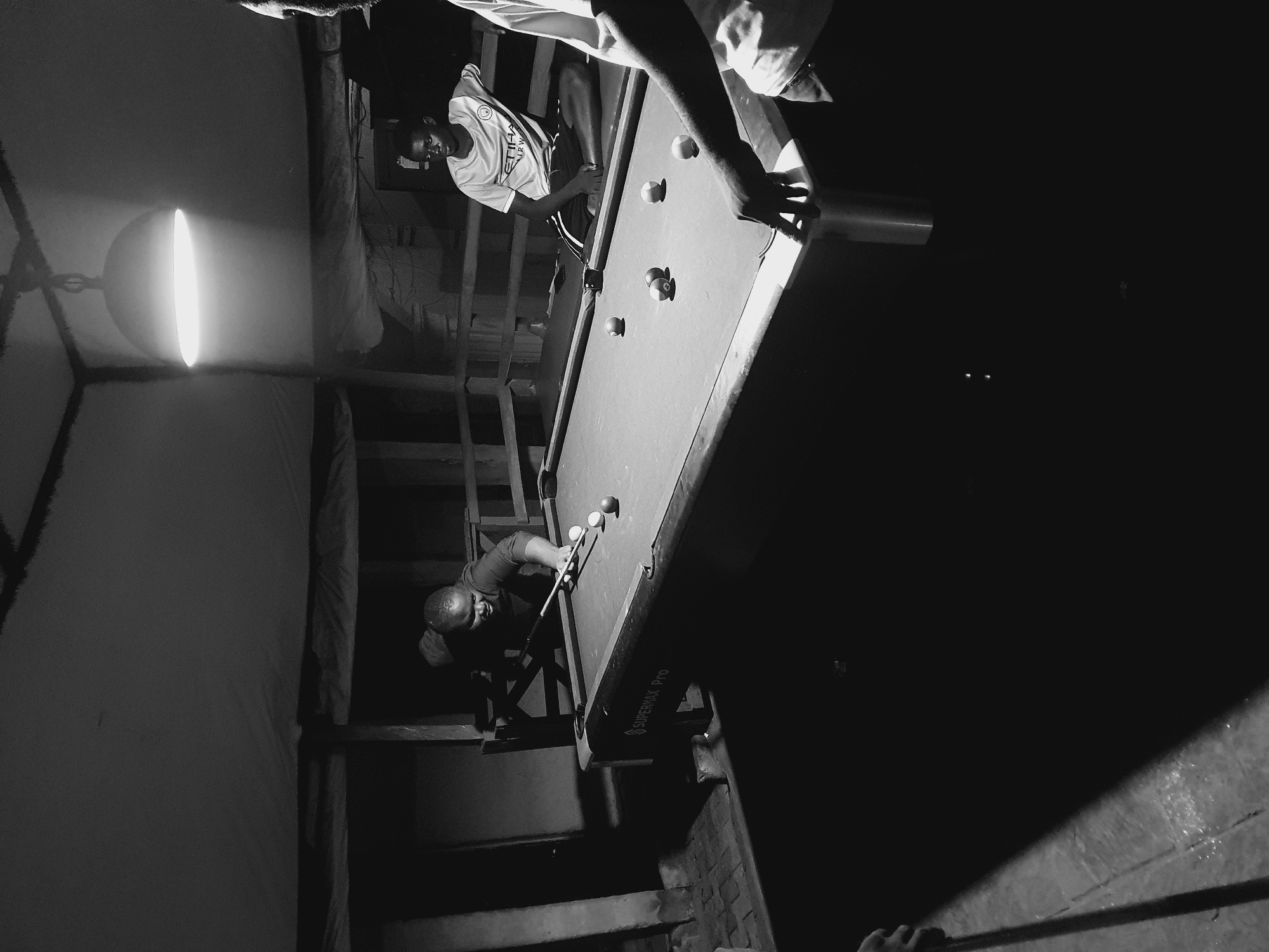 This is a black and white photograph of a group of people playing billiards in what appears to be an indoor, dimly lit space. The image is rotated, showing the players and the pool table at an unusual angle.
The main subjects are three individuals. One person, wearing a t-shirt and shorts, is actively playing pool, aiming a cue at the balls on the table. Another person, wearing a white t-shirt, stands behind the playing person, observing. A third person's arm is visible reaching towards the pool table, suggesting their involvement in the game. The pool table itself is a prominent object, with several balls scattered on its surface. A bright, elongated light fixture hangs from the ceiling, providing the primary illumination for the scene.
The setting seems to be a rustic or unfinished indoor space, possibly a community center, a makeshift recreational area, or even a residential interior not designed for professional use. Wooden beams and planks are visible in the background, suggesting a construction or renovation stage, or simply a utilitarian aesthetic. The floor appears to be made of concrete or rough material.
The activity taking place is a game of billiards, characterized by the pool table, cue stick, and balls. The mood seems casual and focused, with the players engaged in the game. The lighting creates strong contrasts, casting shadows and highlighting certain areas, contributing to a somewhat dramatic or intimate atmosphere. The time of day is not explicitly discernible, but the artificial lighting suggests it could be evening or nighttime, or an interior space where natural light is limited. There are no clear indicators of weather.
There is visible text on the side of the pool table that reads "SUPERMAX PRO". The location context is provided as Lokoja, Nigeria.