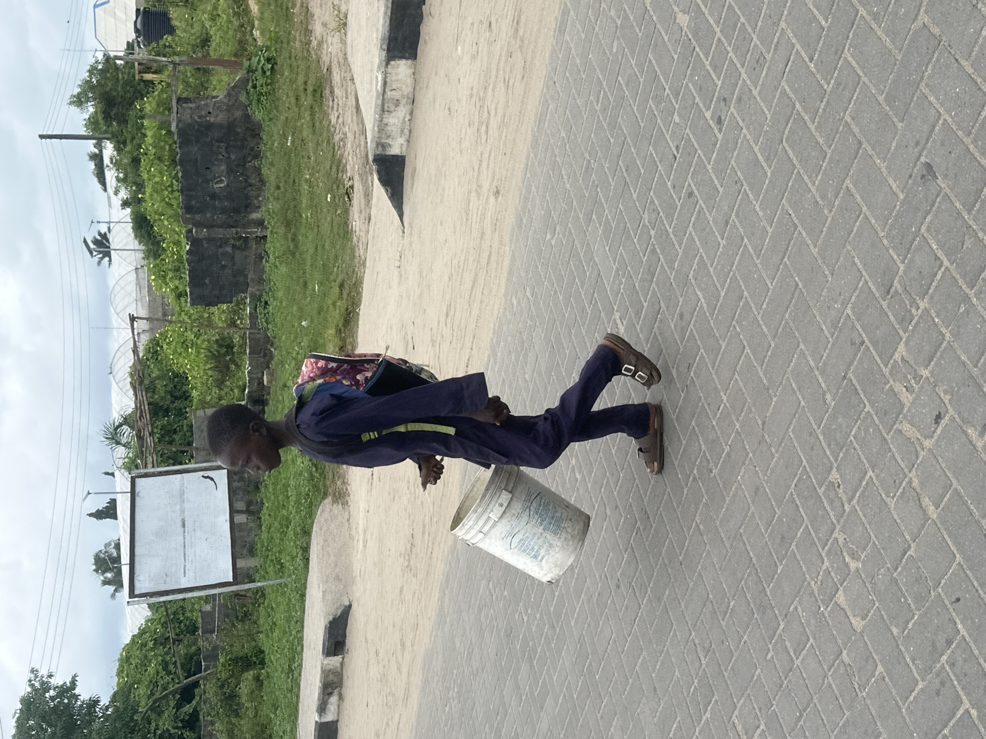 A young boy in a dark blue uniform and sandals walks along a paved path, carrying a white bucket in his right hand and a backpack on his shoulders. The scene takes place in Badagry, Nigeria, with a sandy area and a sidewalk bordering the path. In the background, a white board with text is visible, along with lush green vegetation and structures that appear to be greenhouses. The sky is overcast.