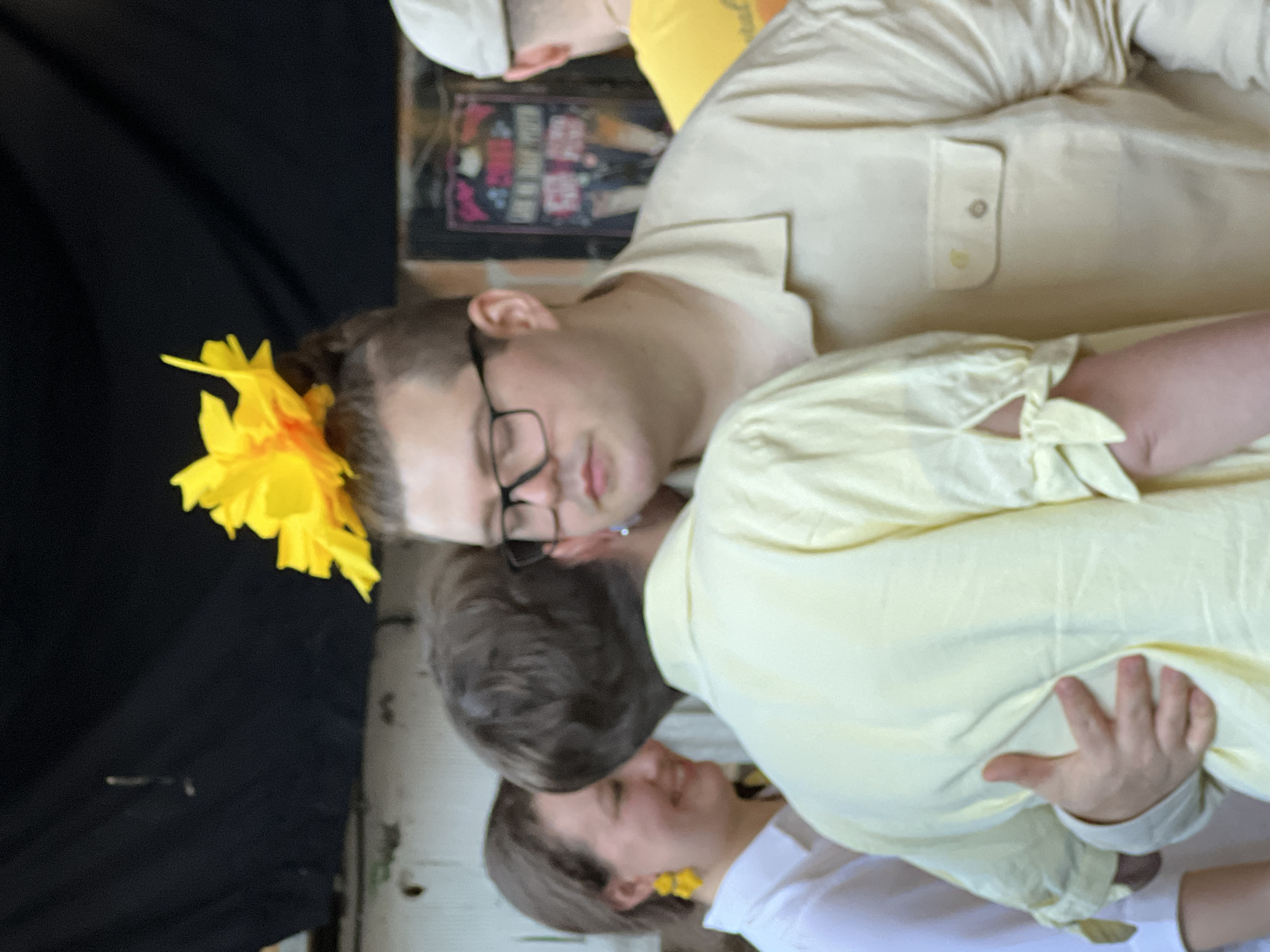 Three people are embracing. One person has a yellow flower in their hair. All are wearing yellow clothing. They appear to be in Belgrade, Serbia, possibly at a celebration.