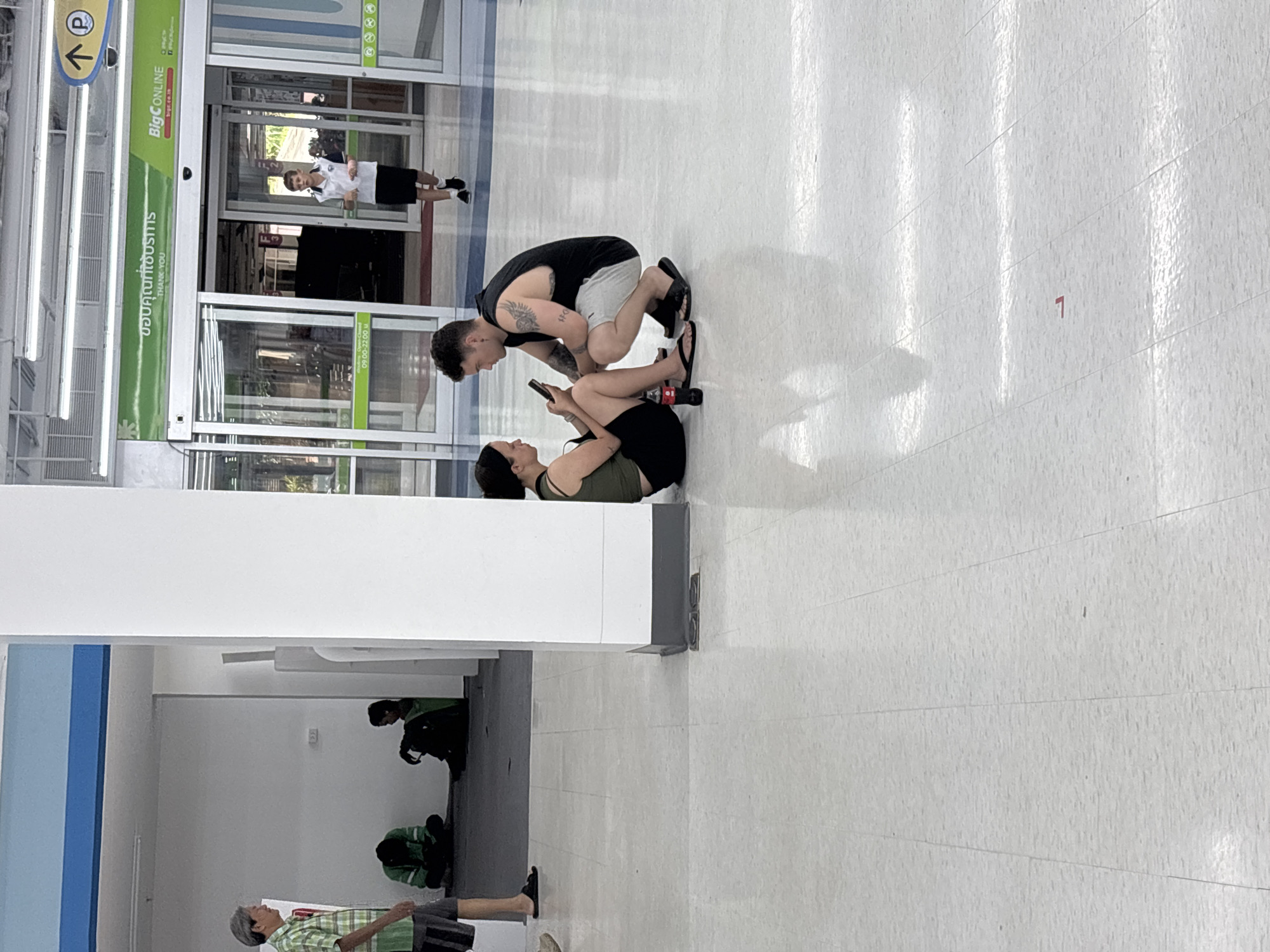 In Baan Bo Phut, Thailand, a young couple sits on the tiled floor in front of a white pillar, both looking at a smartphone held by the woman. She is wearing a dark green tank top and black shorts, with black flip-flops. The man, with tattoos on his arm, is wearing a black sleeveless shirt and light grey shorts, also in flip-flops. A bottle of Coca-Cola and a red soda bottle are on the floor near them. In the background, two individuals are seated on the floor by a white wall. To the right, an employee in a white uniform stands near the entrance of a store with automatic glass doors, which has a green sign reading "BigC ONLINE". An older man in a green and white checkered shirt and shorts stands to the left, facing away from the camera. The environment appears to be an indoor public space, likely a commercial area.