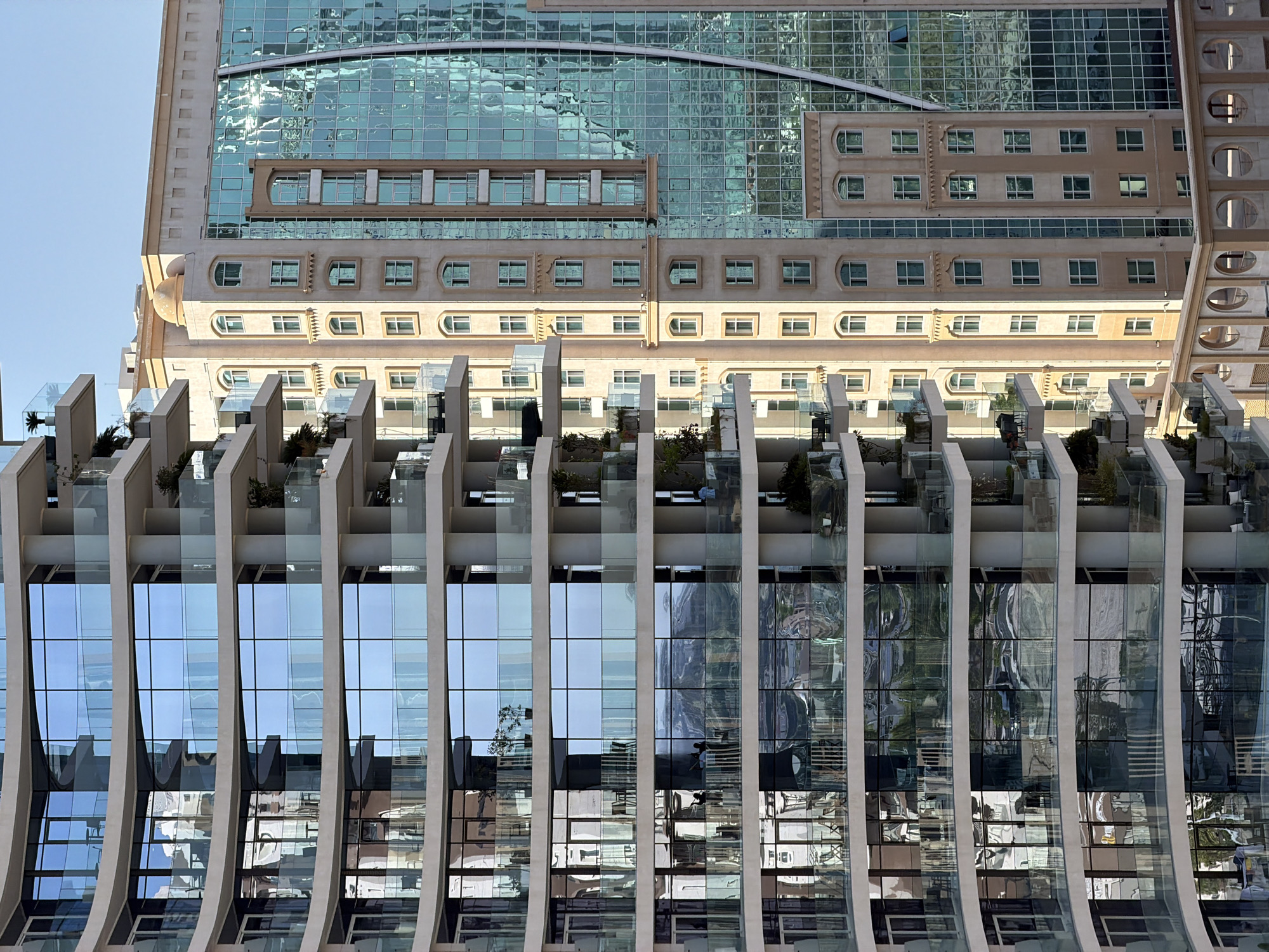 The image displays a detailed view of two distinct architectural structures in an urban setting. On the left, a modern high-rise building features a curvilinear design with multiple levels of balconies equipped with transparent glass railings and light-colored concrete supports. Each level presents large, floor-to-ceiling glass windows that reflect a bright, clear sky, distant buildings, and faint interior details including furniture and potted plants. Some reflections are distorted.
Adjacent to and slightly behind the modern building, a taller structure with a beige, traditional facade stands. This building incorporates multiple vertical sections, some with arched windows and others with rectangular windows arranged in a grid pattern. Decorative elements, such as stepped cornices and possibly corbels, are visible along its upper edge and between floors. A small, light-colored dome crowns a section of this traditional building at the top. A significant portion of this traditional building's right side is covered by a large, blue-green reflective glass facade, creating a stark contrast in materials and style. This glass surface reflects sunlight intensely and shows distorted images of surrounding structures. Further to the right and slightly recessed, another building section with a darker glass facade is visible.
The scene is illuminated by strong daylight, evident from the bright reflections on the glass surfaces and the clear blue sky above. No direct human action or interaction is prominently featured, though faint figures appear in some window reflections. The image captures a blend of contemporary and traditional architectural styles within an urban landscape. The location is an unknown city.