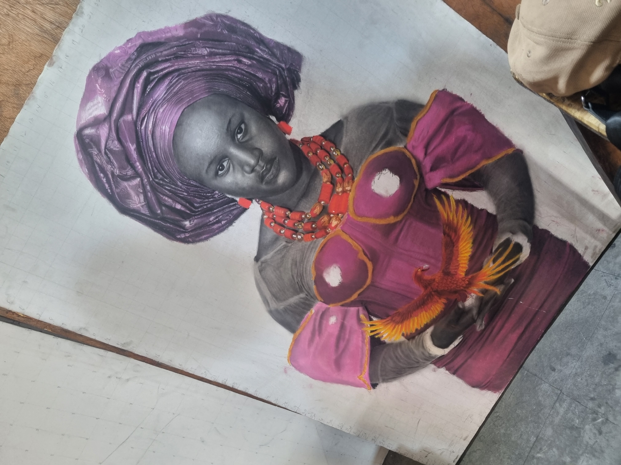A partly finished drawing depicts a woman with dark skin, wearing a violet head wrap and a red beaded necklace and earrings. She is holding a vibrant orange and yellow bird. The woman's face and upper body are rendered in greyscale, while her clothing is colored in shades of pink and red. The drawing is on a piece of paper with grid lines. The surrounding environment shows a wooden table surface and, partially visible, a beige hat. This location is likely a studio or workspace in Onitsha, Nigeria.