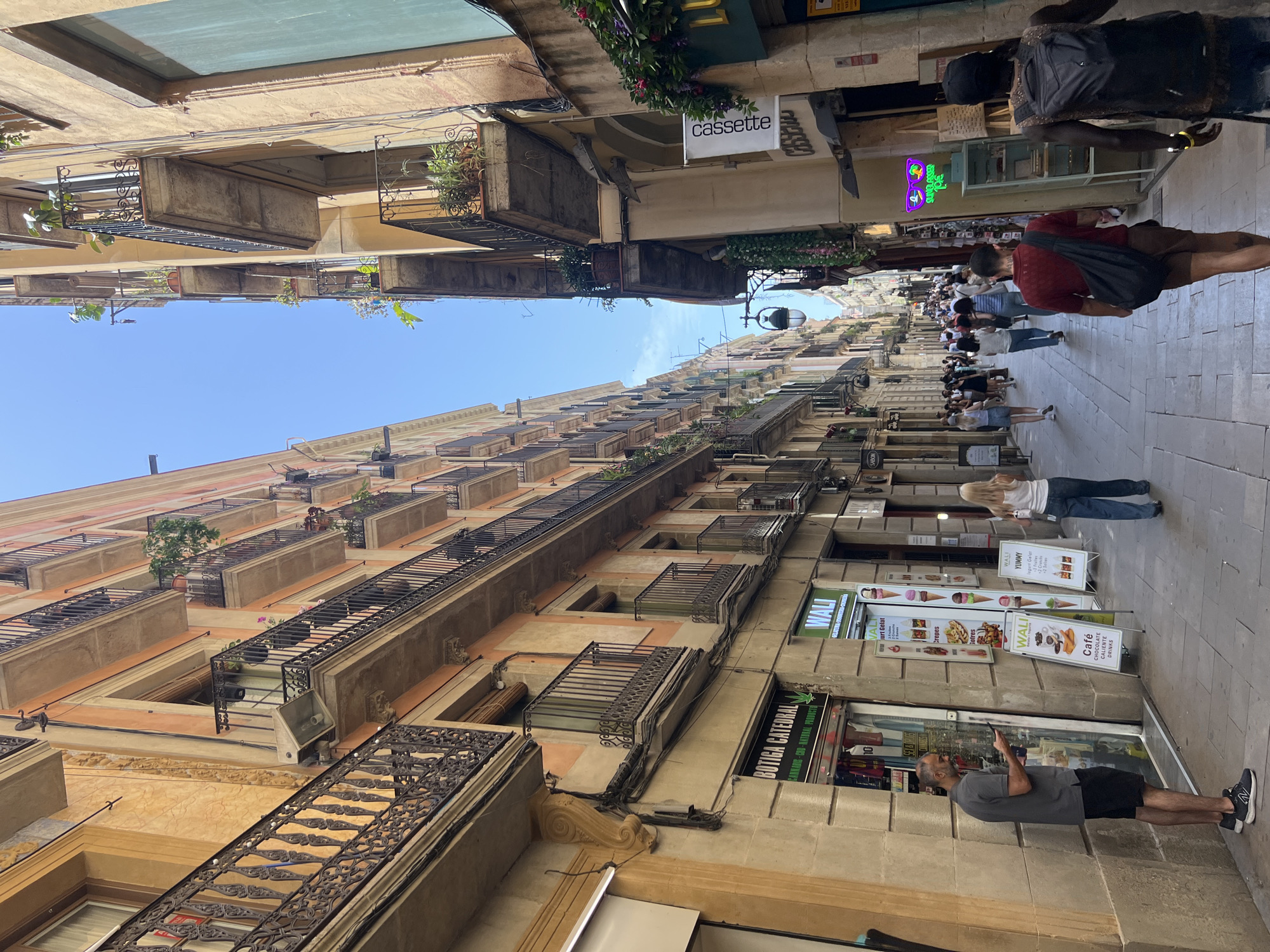 The image depicts a narrow, pedestrianized street in Barcelona, Spain, with buildings lining both sides. The architecture features traditional European style, with balconies adorned with wrought iron railings and planters overflowing with greenery. The street is paved with cobblestones, and several shops are visible at ground level, displaying their wares and advertisements.
In the foreground, a man in a grey t-shirt and black shorts stands near a shop window, looking at his mobile phone. To his right, a woman in a white tank top and blue jeans walks away from the camera. Further down the street, a crowd of people strolls along, suggesting a bustling atmosphere. Shops visible include "BOTIGA CATEDRAL," "WALI," and others advertising food and drinks. A neon sign featuring sunglasses is also visible. The sky above is a clear blue, indicating a sunny day. The overall scene conveys a lively urban environment characteristic of a European city.