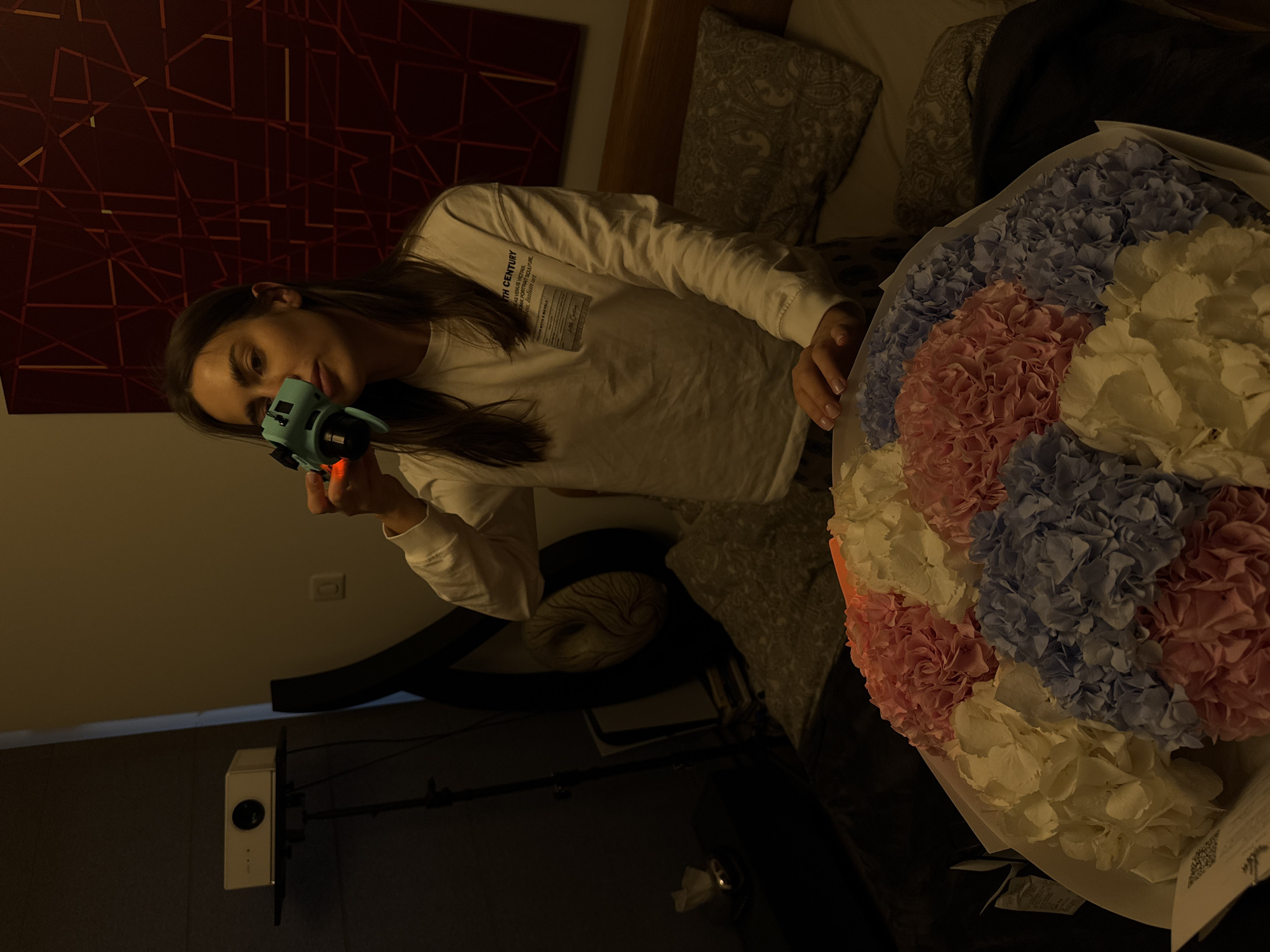 A young woman is lying on a bed, holding a blue toy camera in her right hand, possibly mimicking taking a photo. She is wearing a long-sleeved white shirt with text visible on the chest area. To her right is a bouquet of flowers in shades of white, pink, and blue. The setting appears to be indoors, with a painting on the wall behind the bed and pillows with patterned covers. A device possibly a projector or similar electronic appliance is mounted on the wall in the lower left part of the frame. The exact location is not determinable from the image alone.