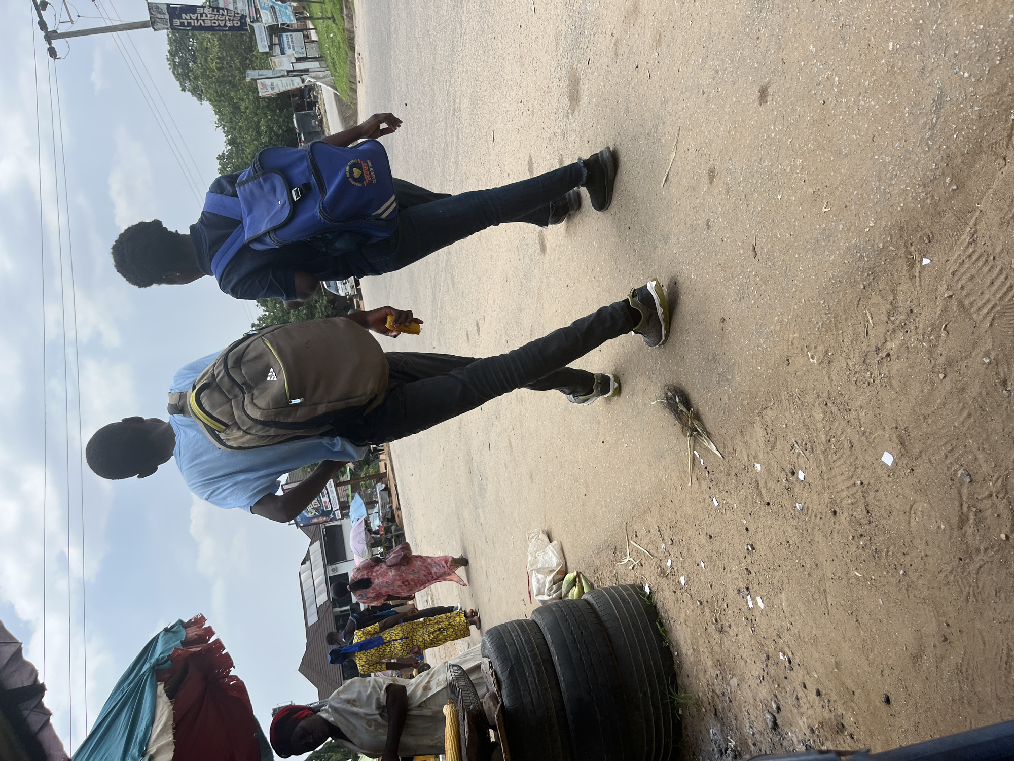 This image captures an outdoor scene in Abuja, Nigeria, likely during the daytime. Two young men are walking along a dusty road. The man in the foreground wears a light blue shirt and a brown backpack, while the man behind him sports a dark shirt and a blue backpack. Both are dressed in dark jeans and sneakers. The man in front is holding a yellow object, possibly food.
The setting appears to be a street market or a busy roadside area. In the background, there are shops and signs, one of which reads "GRACEVILLE CHRISTIAN CENTRE." Several people are visible near the shops, some appearing to be vendors displaying goods such as fabrics and produce like corn. Stacks of tires are also present on the side of the road, along with scattered debris, suggesting a somewhat unkempt environment. The sky is partly cloudy, indicating moderate weather conditions. The overall mood seems casual, with the individuals walking without any apparent urgency.