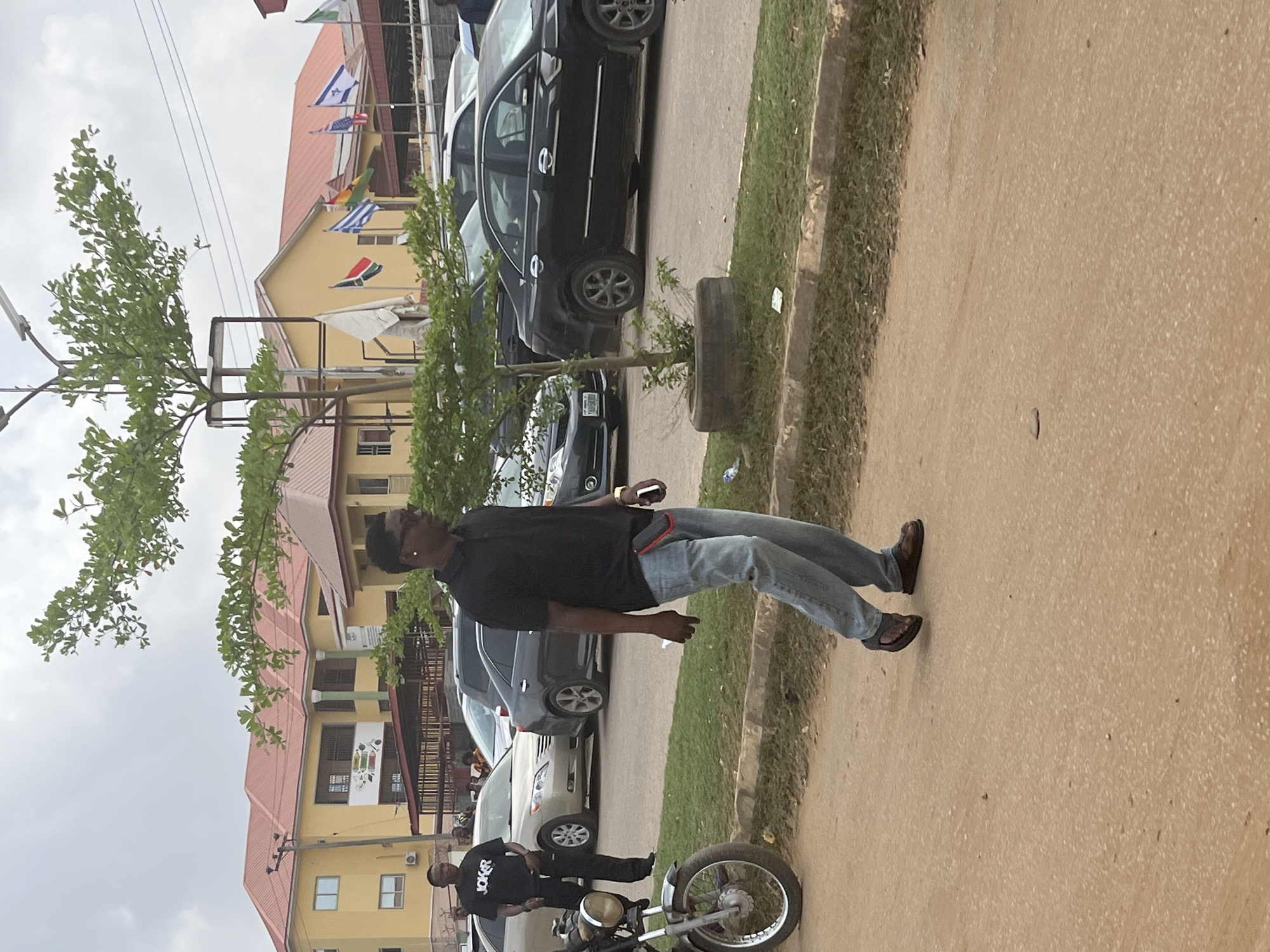 The image depicts a street scene in Abuja, Nigeria, with a man in a black polo shirt and jeans walking on the sidewalk. He holds a phone. Various cars are parked along the street, with a motorcycle near the lower edge of the frame. Another man is standing next to the motorcycle wearing black. In the background, a building is visible, adorned with flags from various countries. Green vegetation grows near the sidewalk. The day seems overcast.